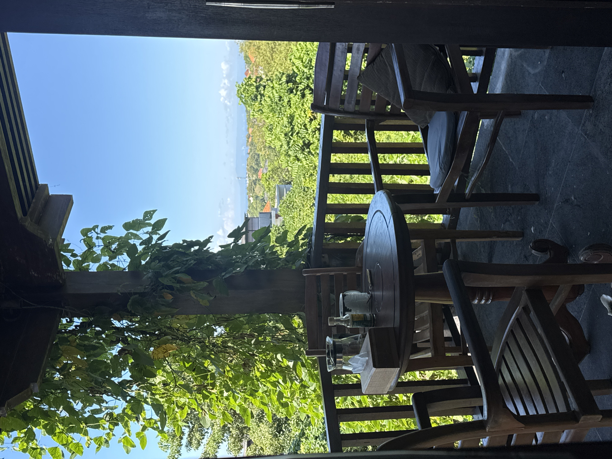 The image shows a view from a covered outdoor patio or balcony. The location is implied to be Kuta Selatan, Indonesia. Wooden chairs and a round wooden table are visible on the patio. The table holds a bottle, a glass, and a tissue box. The patio has a wooden railing. Beyond the railing, there is a lush green landscape with trees and glimpses of buildings. The sky is clear blue. Vines and foliage grow around the patio's support beams, partially obscuring the view. The perspective is from inside, looking out. The focus is on the patio furniture and the view.