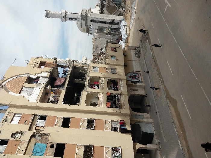 The image depicts a street scene in Khan Yunis, Palestinian Territory, characterized by significant structural damage to buildings. A prominent multi-story residential building, light brown in color, occupies the left side of the frame. It exhibits extensive damage, including numerous shattered windows, exposed interiors, missing wall sections, and pockmarks on its facade. Blue and red fabrics cover some window openings, and laundry hangs from several damaged balconies. An arched decorative element is visible on an upper floor. To the right and in the background, a heavily damaged mosque features a partially collapsed dome and a scarred but largely standing minaret.
In the foreground, a paved two-lane road with white lane markings, including a turn arrow, runs horizontally. Adjacent to the building, a gray paved sidewalk is littered with rubble and debris. Several individuals are present: two adults in dark clothing walk along the road in opposite directions, one adult sits on the sidewalk, and at least two children are visible near the ground floor of the damaged building, one near a doorway covered by a black tarp. Part of another individual, possibly wearing a headscarf, is visible in the bottom left corner of the frame. The sky is partially cloudy.