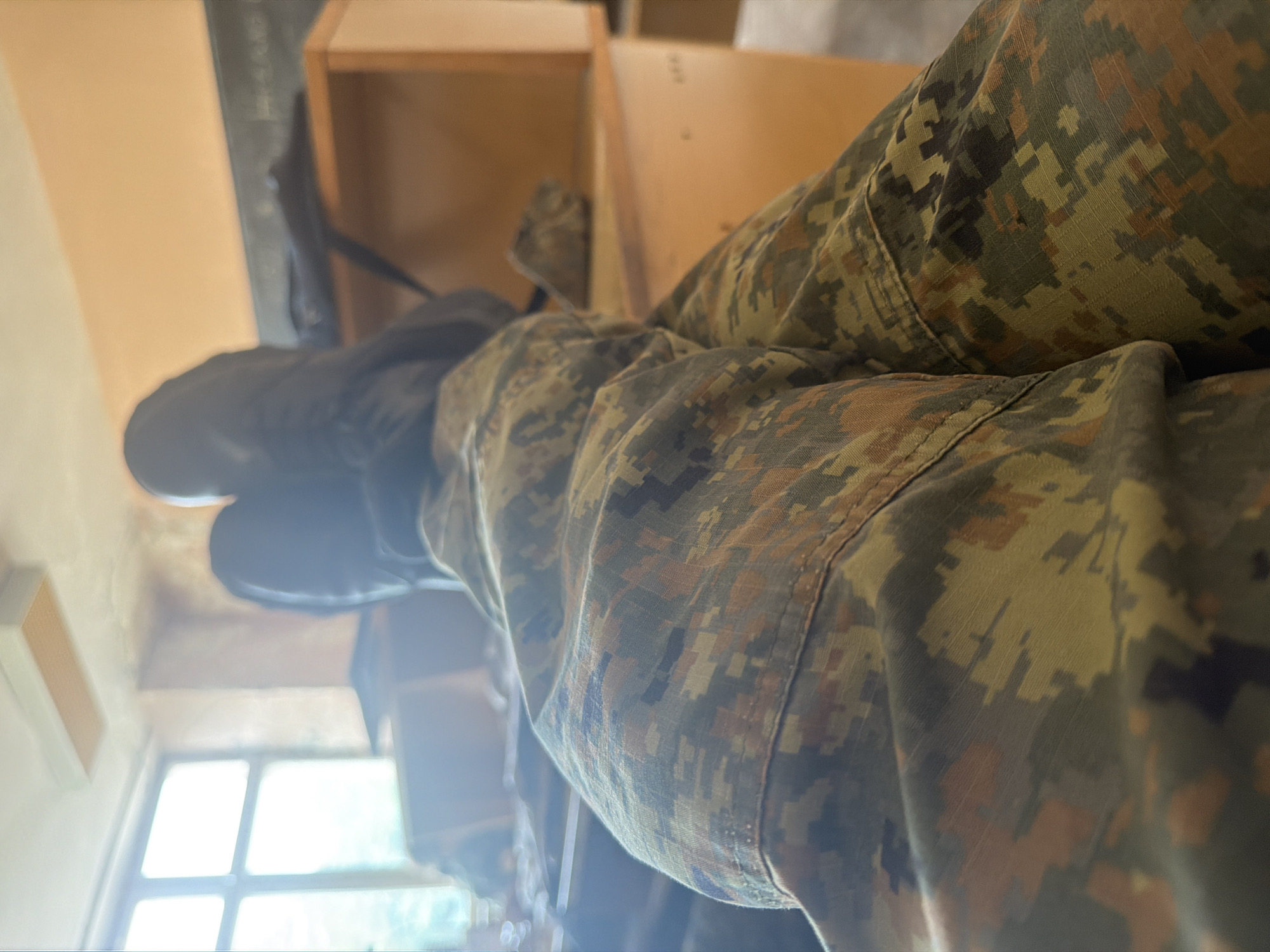 A close-up shot focuses on a person's legs clad in camouflage print trousers. The person is seated. In the background, a window with a light source. The location and city are impossible to be verified.