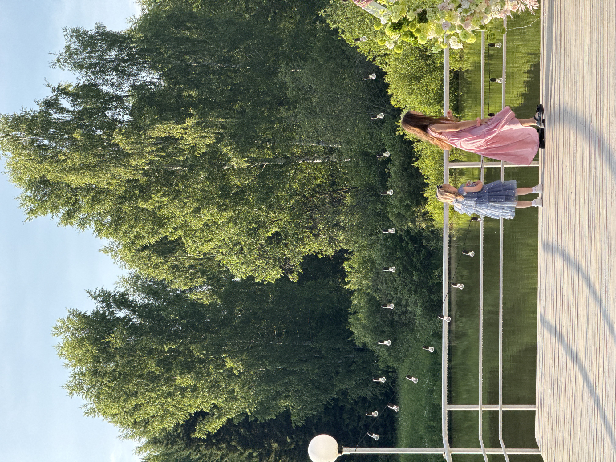 The image depicts two young girls on a wooden pier near a body of water, surrounded by lush green trees under a clear blue sky. The girl on the left is wearing a blue dress with white polka dots and white shoes. The girl on the right is wearing a pink dress and dark shoes. They are both facing away from the camera, towards the water. A white railing runs along the edge of the pier. String lights are visible hanging above the railing. In the background, dense foliage and trees create a natural setting. The lighting suggests it is daytime, possibly late afternoon, with the sun casting shadows on the pier. The location is identified as Votkinsk, Russia.