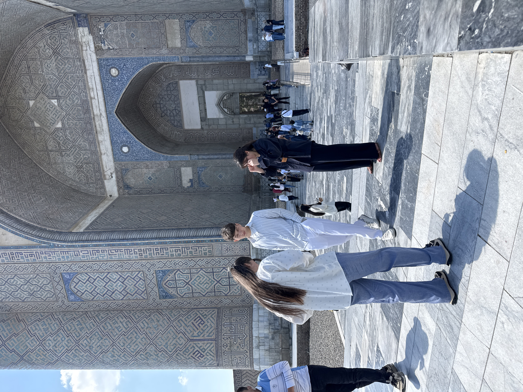 The image shows three tourists standing in front of a large building in Samarkand, Uzbekistan. The building has intricate blue and white tilework, and appears to be an Islamic religious site, potentially a mosque or a mausoleum. Other visitors are further inside the building. The tourists are oriented in parallel to the facade of the building. The floor is covered with large grey rectangular slabs. The sky is partially visible in the lower left.