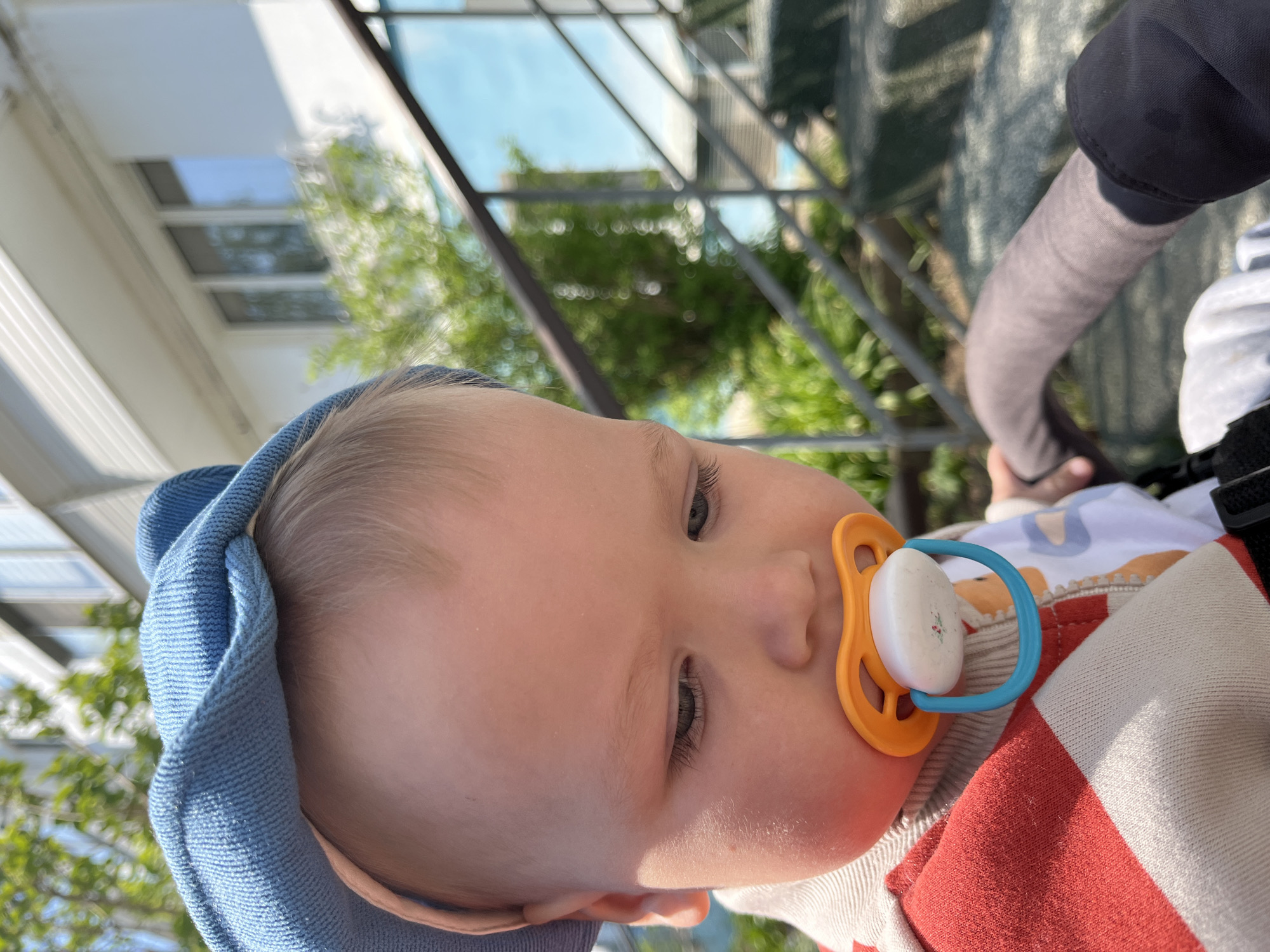 A baby with a blue head covering and an orange and blue pacifier in its mouth is the central subject of the image. The baby is lying down in what appears to be a stroller or similar conveyance, as indicated by a gray-covered bar visible to the right. The baby is wearing a red and white striped garment over a patterned shirt. The background shows glimpses of building facades, greenery suggesting trees, and parts of what may be an architectural frame structure. The location might be in Chaikovsky, Russia, though the photograph does not provide any specific geotags or landmarks to confirm the location. There are no explicit actions or interactions visible other than the baby's presence with the pacifier in the described setting.