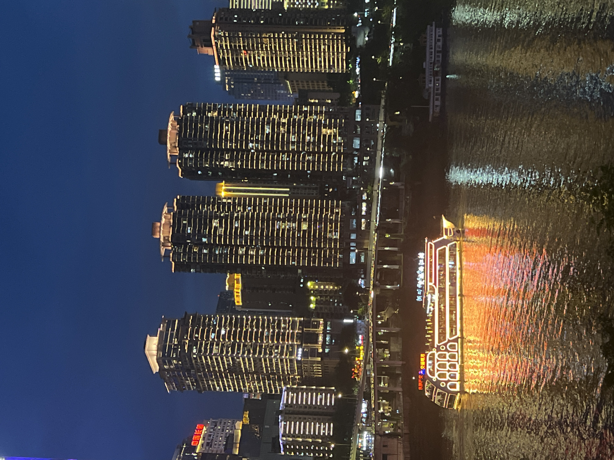 This is a nighttime photograph taken from a high vantage point, overlooking a city skyline and a body of water. The scene is dominated by towering, modern apartment buildings illuminated with countless warm lights, creating a striking contrast against the deep blue twilight sky. The buildings are arranged in a row along the waterfront, with their facades featuring repeating patterns of windows and vertical lighting elements.
In the foreground, a body of water reflects the lights from the buildings and a brightly lit tour boat. The boat, with its distinctive design and glowing exterior lights in yellow, orange, and red, is a prominent feature, its reflection shimmering on the rippling surface of the water. The water's surface is textured with the movement, causing the reflections to appear fragmented and dynamic.
The overall atmosphere is one of urban vibrancy and nighttime activity. The scene suggests a city in China, specifically near JiangbeiCheng Street, with a lively waterfront that attracts nighttime visitors. The time of day is clearly evening or night, as evidenced by the dark sky and the extensive artificial lighting. The weather appears to be clear, allowing for sharp reflections on the water. No specific human activities are clearly depicted, but the illuminated buildings and the tour boat imply a populated and active urban environment. Visible text on the tour boat includes Chinese characters.