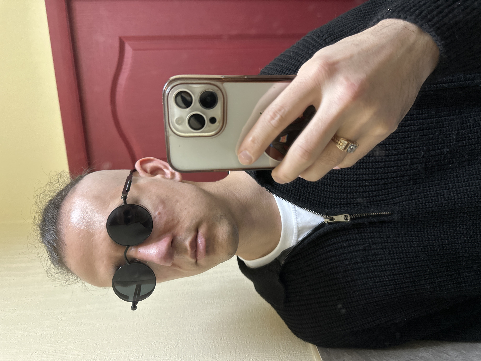 A male individual is depicted in a self-portrait (selfie) taken indoors. The scene is located in Saratov, Russia. The male, an adult with light skin tone, wears dark, round-frame sunglasses with small embellishments on the outer edges. His hair is short on the sides, longer and styled upwards on top, with visible grey strands. He is dressed in a white crew-neck t-shirt layered under a black, ribbed-knit quarter-zip sweater with a silver zipper. His right hand holds a light-colored smartphone with a visible triple camera array and a protective case, positioning it to capture the image. A gold-toned ring with a rectangular or square stone is present on the ring finger of his right hand. The background consists of a textured, light-colored (cream or beige) wall on the left and a red-painted wooden door with a panel design on the right. The top portion of the background above the door is a slightly darker yellow or cream color. The lighting appears to be even, illuminating the subject from the front.