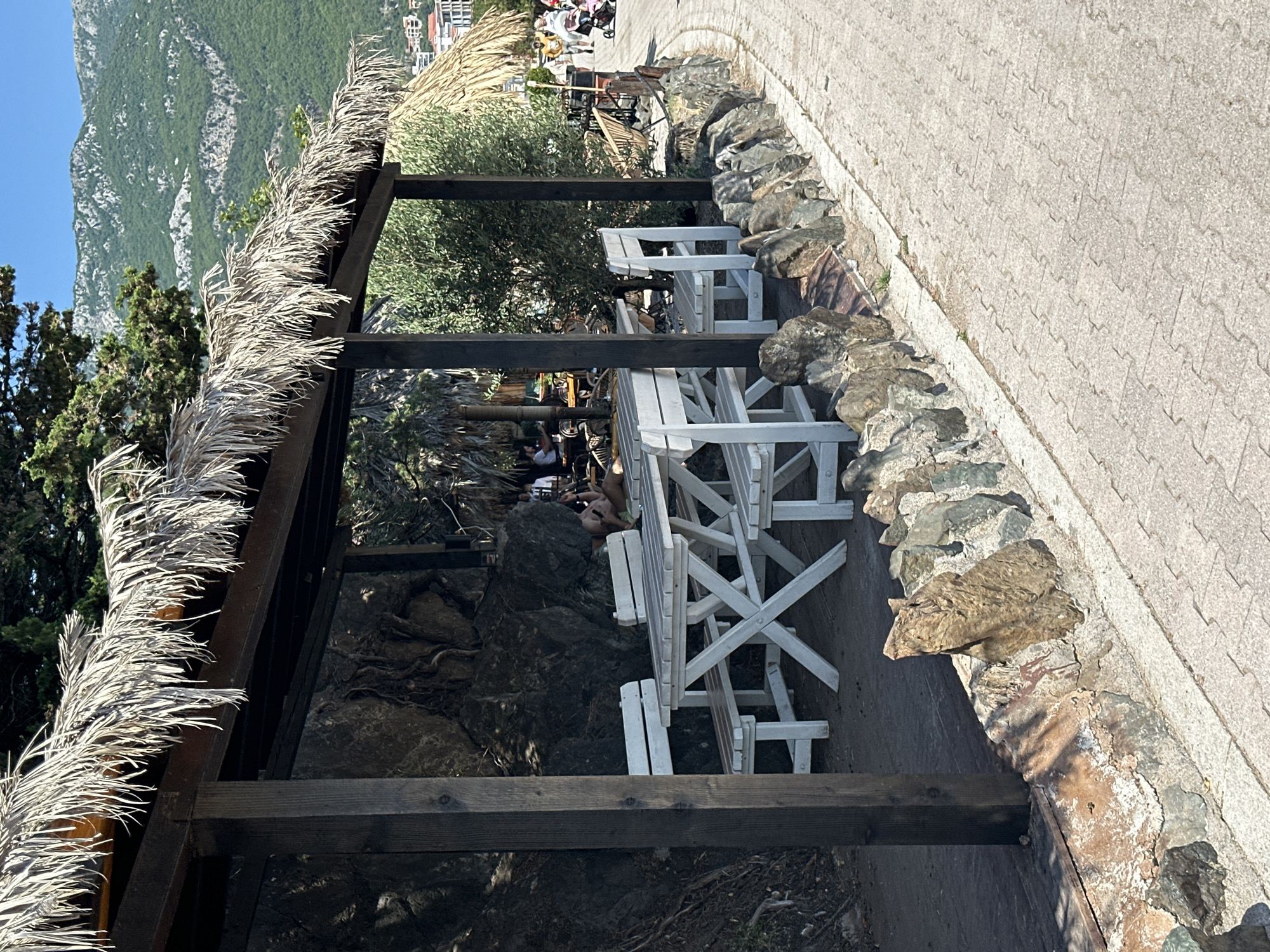 This is an outdoor scene in Rafailovići, Montenegro, featuring a rustic outdoor seating area. A thatched roof structure provides shade over a white wooden picnic table and benches. Behind the table, a large rock face is visible, partially covered with greenery. To the right of the seating area, a paved pathway curves away, bordered by a low stone wall. In the background, a lush green mountainside rises, with some buildings visible near its base. Further down the pathway, other patrons and a stroller are seen, suggesting a public promenade or beach area. The overall atmosphere is relaxed and summery.