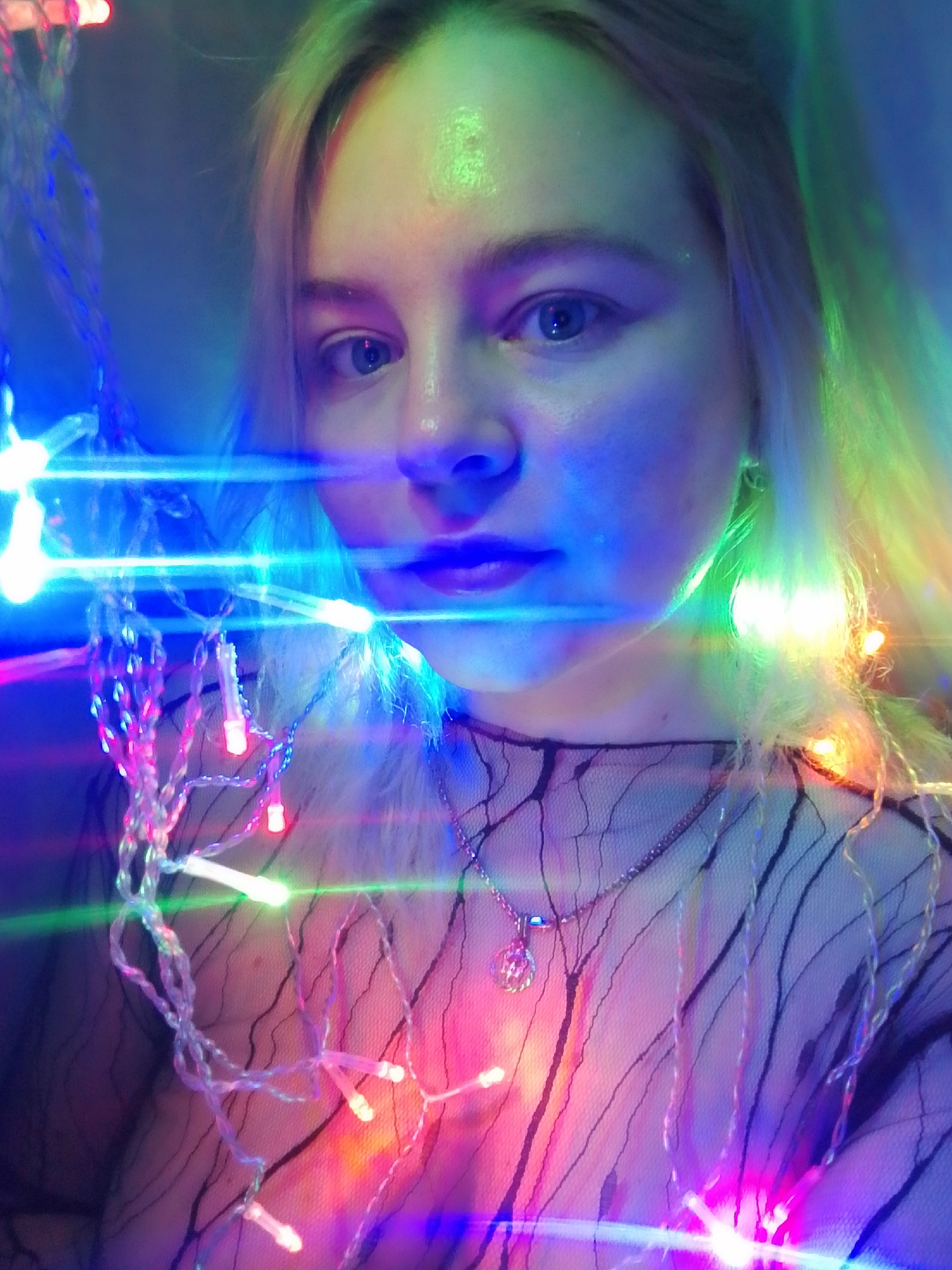 A fair-skinned person with blonde hair is centered in the frame, their face illuminated by multicolored string lights. The lights are draped around the person, obscuring parts of their face and body. The person is wearing a sheer, dark-colored top with a pattern resembling a web or netting and a silver necklace with a pendant. No specific location in Izhevsk, Russia is identifiable from the image.