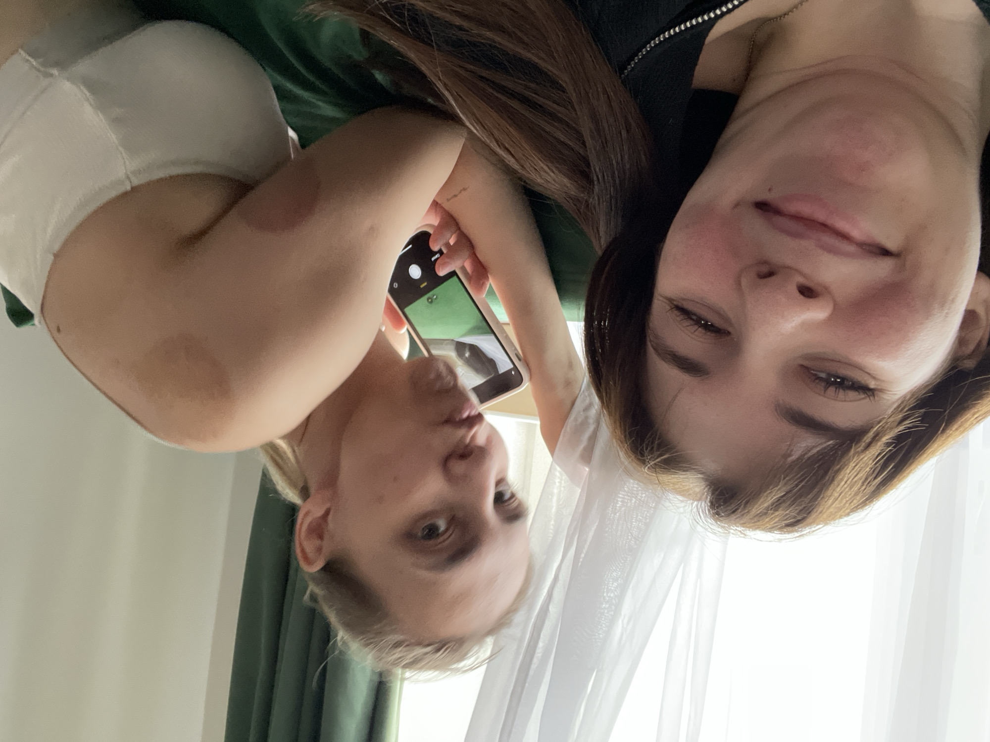 Two women are lying on a surface, possibly a bed or couch, taking a selfie with a smartphone. One woman has blonde hair and is pouting at the camera, while the other has brown hair and is smiling slightly. The woman taking the photo is wearing a white top, and the other woman is wearing a black top. There are dark red marks on the arm of the woman taking the picture. The background includes green and white curtains. It's impossible to confirm the location in Chaikovsky, Russia, from this single image.