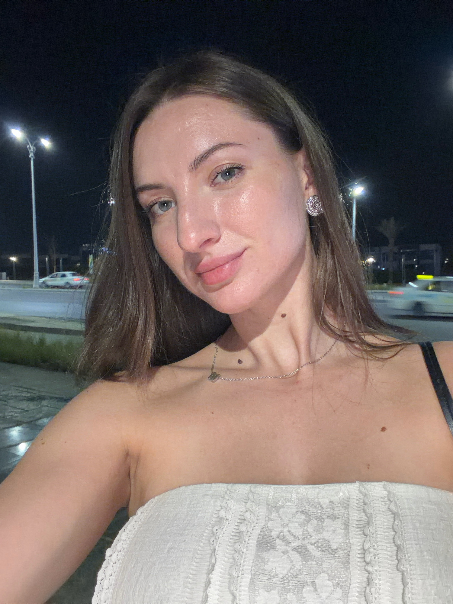 A medium shot selfie taken at night in Sharm El Sheikh, Egypt shows a woman with long, brown hair and blue eyes. She is wearing a white strapless top and a silver necklace. Her skin appears dewy and illuminated by the artificial lights, possibly streetlights or car headlights, which are visible in the background along with palm trees and a road with passing cars. She has a mole on her neck and another on her upper chest. A decorative, rose-shaped earring is visible on her right ear. The background suggests an outdoor urban setting at night, possibly near a street or promenade.