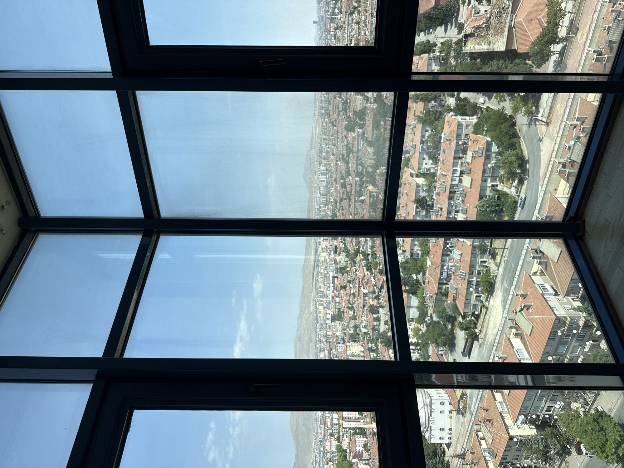 The image shows a panoramic view of the city of Konya, Turkey, taken from a high-rise building through a multi-pane window. The foreground features dark blue window frames, creating a grid-like effect that overlays the cityscape.
The city itself is characterized by a dense arrangement of buildings with terracotta roofs, interspersed with trees and greenery. The architecture appears to be a mix of residential blocks and what seem to be public buildings, indicated by a larger, lighter-colored structure with a domed roof visible on the left side of the image. Roads and smaller streets wind through the urban landscape, with a few vehicles visible. In the middle ground, the city stretches out towards a hazy horizon, where low, undulating hills form the backdrop. The sky is a clear, pale blue with a few scattered clouds.
The perspective suggests a bright, sunny day. The overall impression is of a sprawling, well-established urban environment. No people are discernable in the image. The scene is static, offering a still glimpse of the city.
