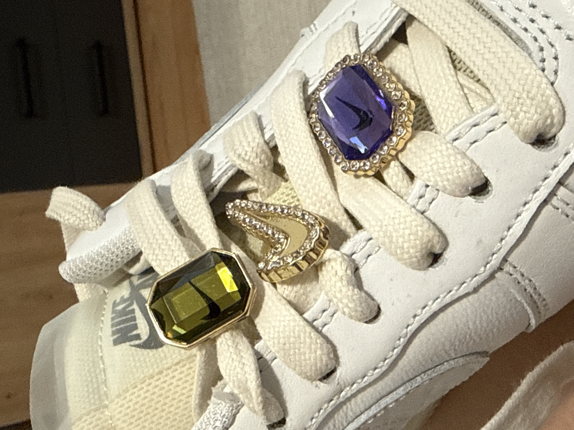 A close-up shot captures the front upper section of a white or off-white Nike sneaker adorned with decorative charms, likely in Hainburg an der Donau, Austria. The sneaker features matching textured shoelaces tied in a crisscross pattern.
Three ornate charms are attached to the shoelaces. The uppermost charm is a large, faceted purple gemstone, possibly amethyst, set in a gold-colored metal frame encrusted with small, clear rhinestones. A distinct black Nike swoosh logo is visible, reflected within the purple gem. Below this, a smaller, gold-colored charm shaped like a 'V' or chevron is also embellished with small, clear rhinestones. The lowest charm is another large, faceted gem, this one an olive green color, possibly peridot, also set in a gold-colored metal frame. A reflection of the black Nike swoosh is also visible within the green gem.
The tongue of the sneaker, partially visible below the charms, displays the word "NIKE" in dark grey lettering, accompanied by its iconic swoosh logo. The leather and fabric textures of the shoe are clearly visible, indicating attention to detail in its construction. The background is softly blurred, showing muted tones of brown and dark grey, suggesting an indoor setting with wooden elements. The focus is entirely on the sneaker and its embellishments, showcasing a personalized or customized footwear item. No people are visible, and the activity depicted is the display of the sneaker. No specific time of day or weather can be inferred.