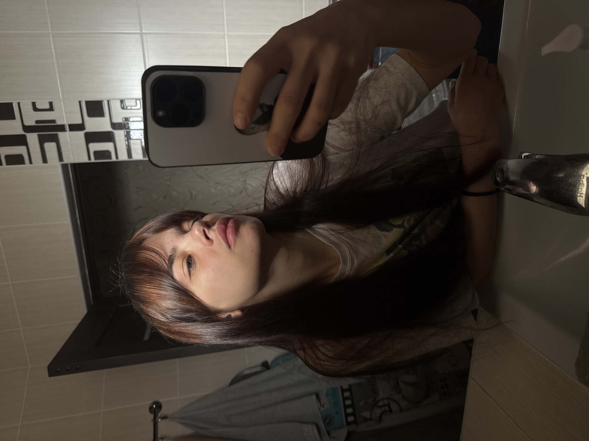 A woman is taking a selfie in a bathroom. The location is potentially in Rostov-on-Don, Russia. The bathroom features tiled walls with a decorative border. The woman is holding a smartphone in her right hand to capture the image. She has long dark hair with reddish-brown highlights and bangs. Her face is visible, and she is looking at the camera. A sink is visible below the phone. The faucet is a metallic color. A towel is hanging in the lower part of the image. The overall lighting is somewhat dim.