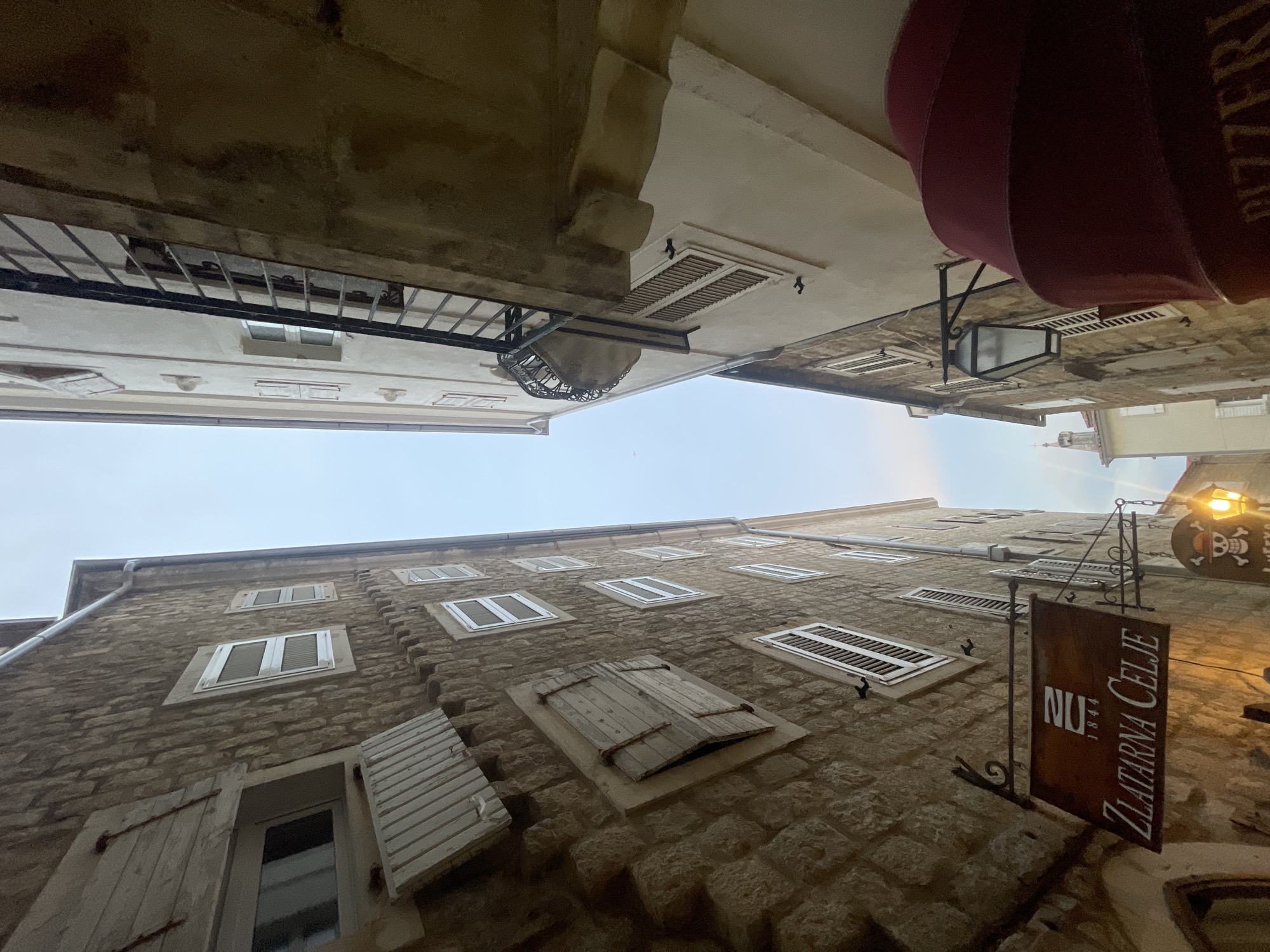 This is an eye-level, wide-angle photograph taken from a low perspective, looking upwards at the facades of buildings lining a narrow street or alley. The buildings appear to be old, constructed with stone blocks, and have multiple windows, some with wooden shutters. The scene is lit by natural light from an overcast sky, and by artificial lights emanating from a lantern and a sign.
The buildings are tightly packed, suggesting an urban or historical setting. The architecture indicates an older style, with details like the stone work, the wooden shutters, and the wrought-iron balcony railing contributing to this impression. The perspective creates a sense of looking up into the sky from a confined space.
One building on the right has a hanging sign with the text "NU ZLATARNA CELJE" and the year "1844". Above this sign is a decorative lantern with a skull and crossbones motif visible on a circular plaque. A dark red awning is also visible extending from an upper floor.
The overall mood is somewhat subdued due to the overcast sky, but the warm glow of the lantern and the illuminated sign add a touch of character and visual interest. The time of day appears to be late afternoon or early evening, given the quality of the light and the illumination of the artificial lights. There are no people visible in the frame. The architecture and the text on the sign provide clues to the location potentially being in or related to Slovenia, given "Celje" is a city there.
