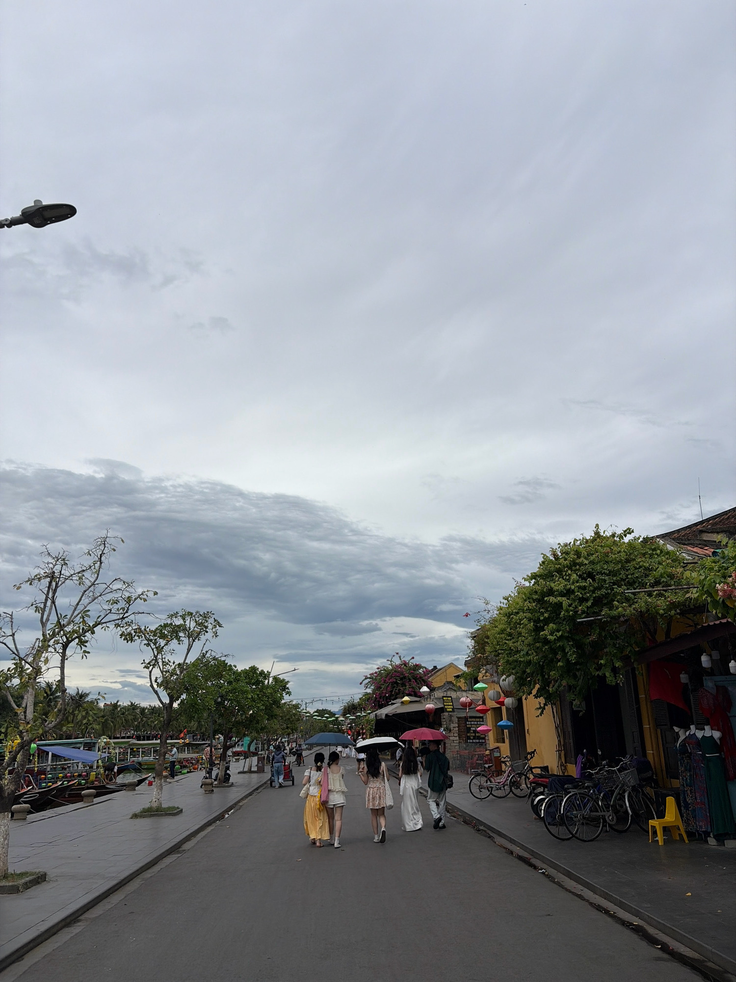 This image depicts a street scene in Hoi An, Vietnam, likely during overcast weather, given the cloudy sky and the presence of umbrellas. On the left side of the street, a canal or river is visible, lined with boats and people. The right side of the street features shops with clothing displayed outside, and several bicycles are parked along the sidewalk. Four individuals, three women and one person in a green jacket, are walking away from the camera down the street. One of the women is carrying a pink umbrella. The architecture suggests a historic district, consistent with Hoi An's Old Town.