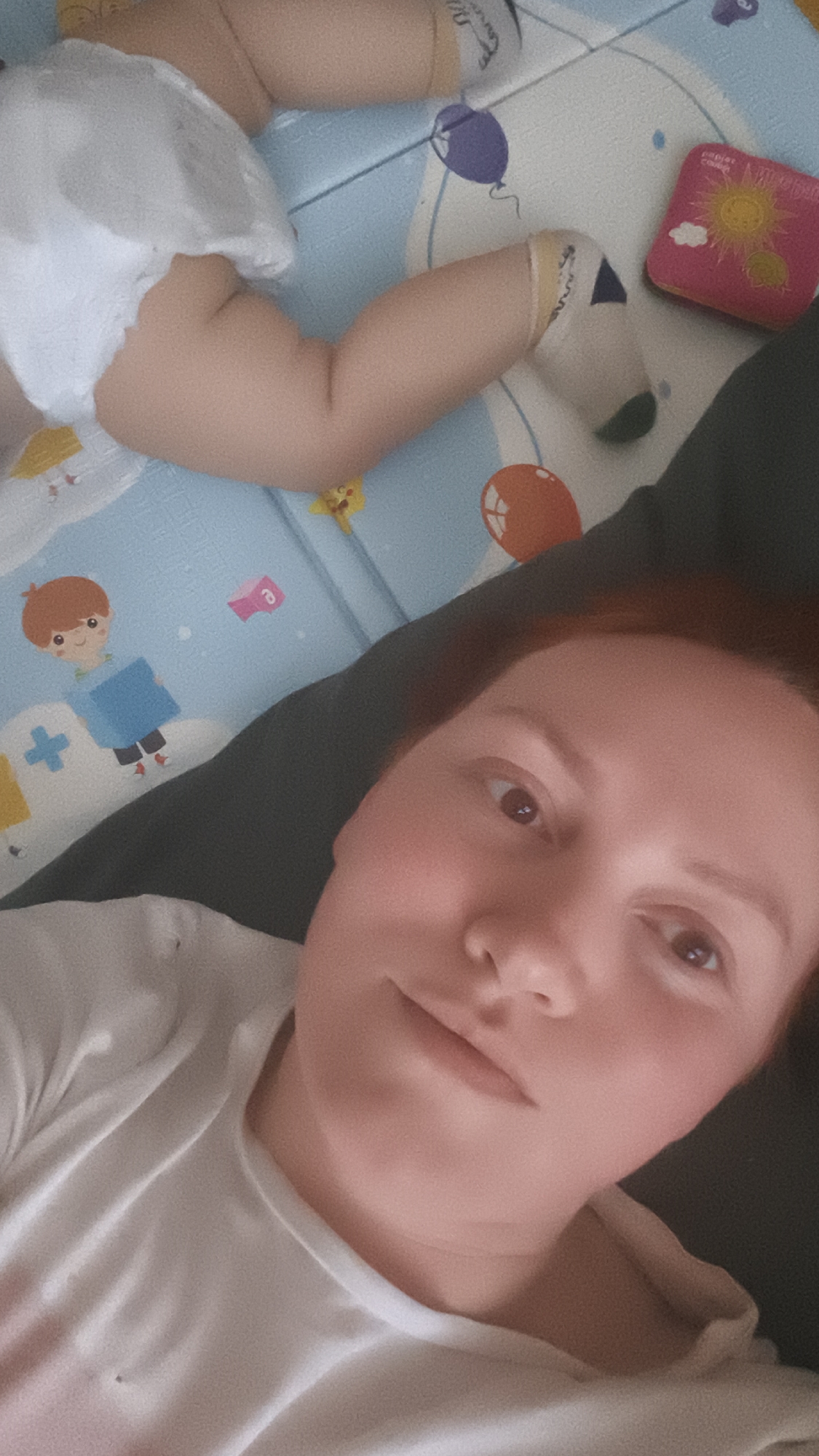 The image shows a woman lying on a dark grey surface, taking a selfie. Parts of a baby are visible on a colorful play mat behind her. The mat has a light blue base with images of balloons, a cartoon boy, and a sun. The baby is wearing a diaper and socks. The overall location appears to be indoors.