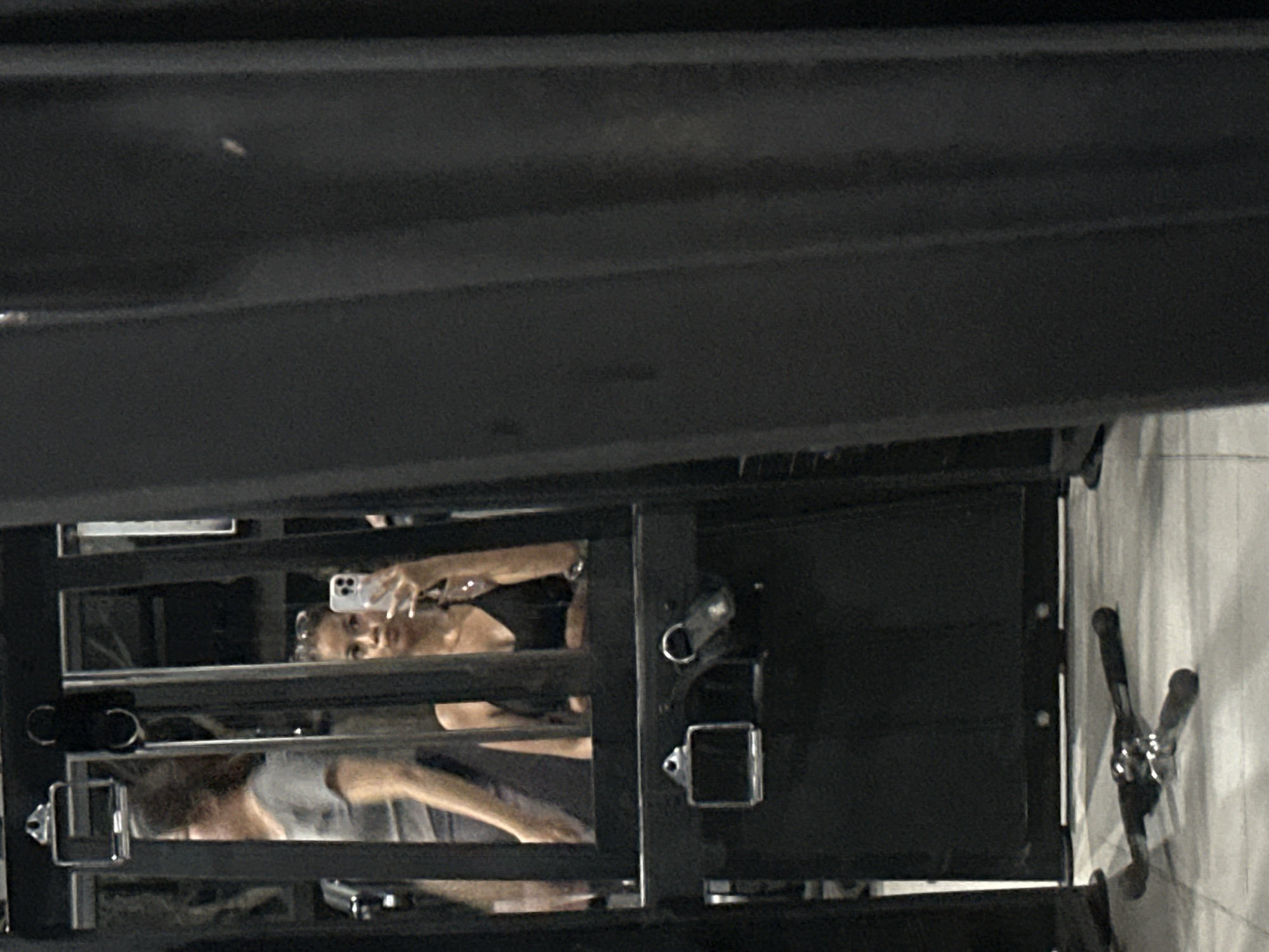 A partially obscured view, looking through the frame of what appears to be metal scaffolding or a theatrical lighting rig. Two individuals are visible through the grid. The individual at the top is holding a smartphone and appearing to take a picture of themselves. The setting may be a backstage or construction area.
There is no evidence of the location of Batubulan, Indonesia.