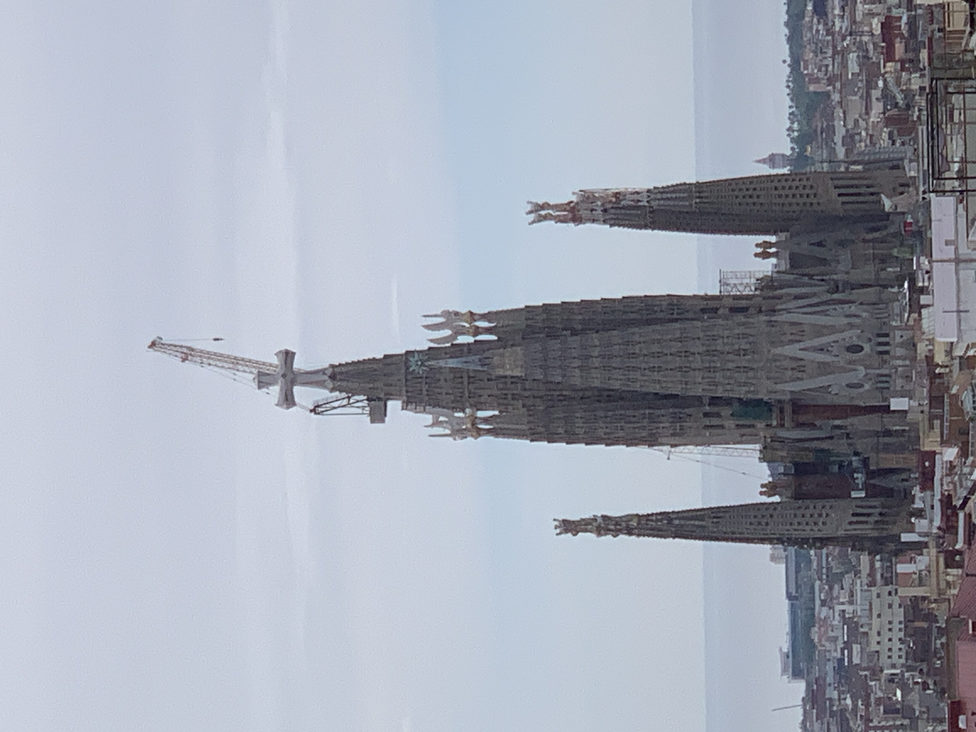 The image shows the Sagrada Familia basilica in Barcelona, Spain. The basilica is under construction, with cranes visible on the main tower. The building is surrounded by the urban landscape of Barcelona, with numerous buildings and houses. The sky is overcast.