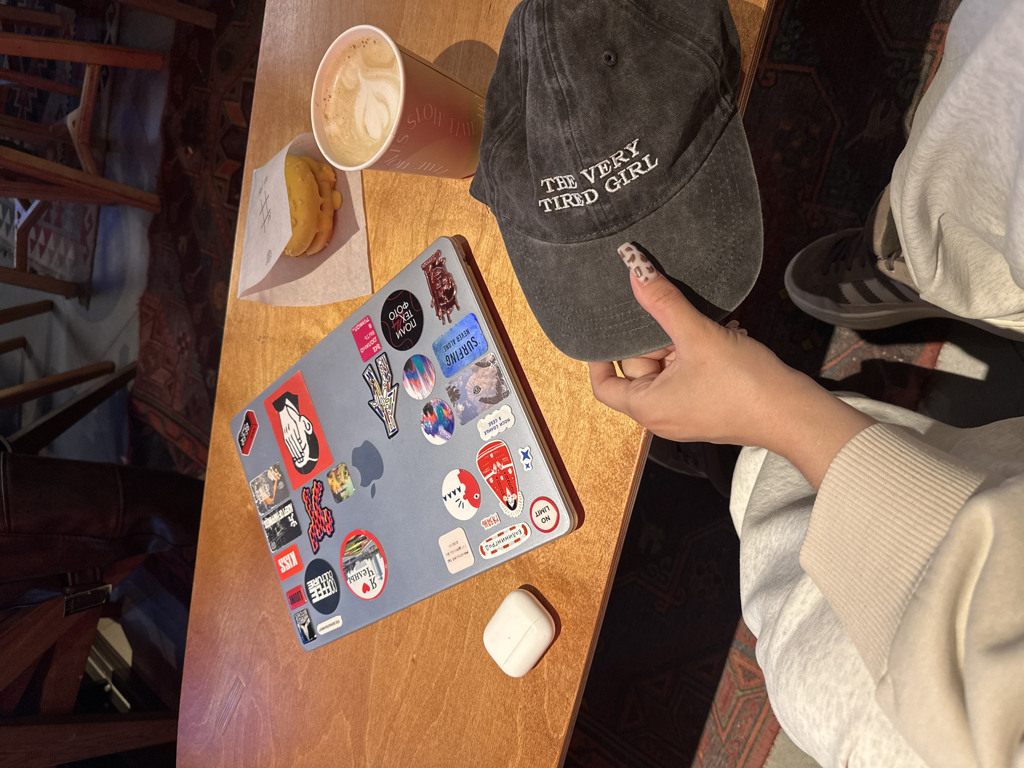 A person wearing a light-colored long-sleeved top is holding a dark gray baseball cap that reads "THE VERY TIRED GIRL" in white text. The person's nails are decorated with a leopard print pattern. The hat is positioned on a light brown wooden table next to a cup of coffee with latte art, a pastry in a white paper bag, and a gray laptop covered with stickers. A pair of white wireless earbuds are also on the table. A corner of a brown bag can be seen next to the laptop. The floor has an ornate rug and what appears to be the legs of furniture. The laptop has several stickers, some with writing and designs. One sticker features a hand graphic with a red background, and others have text in both English and Cyrillic.