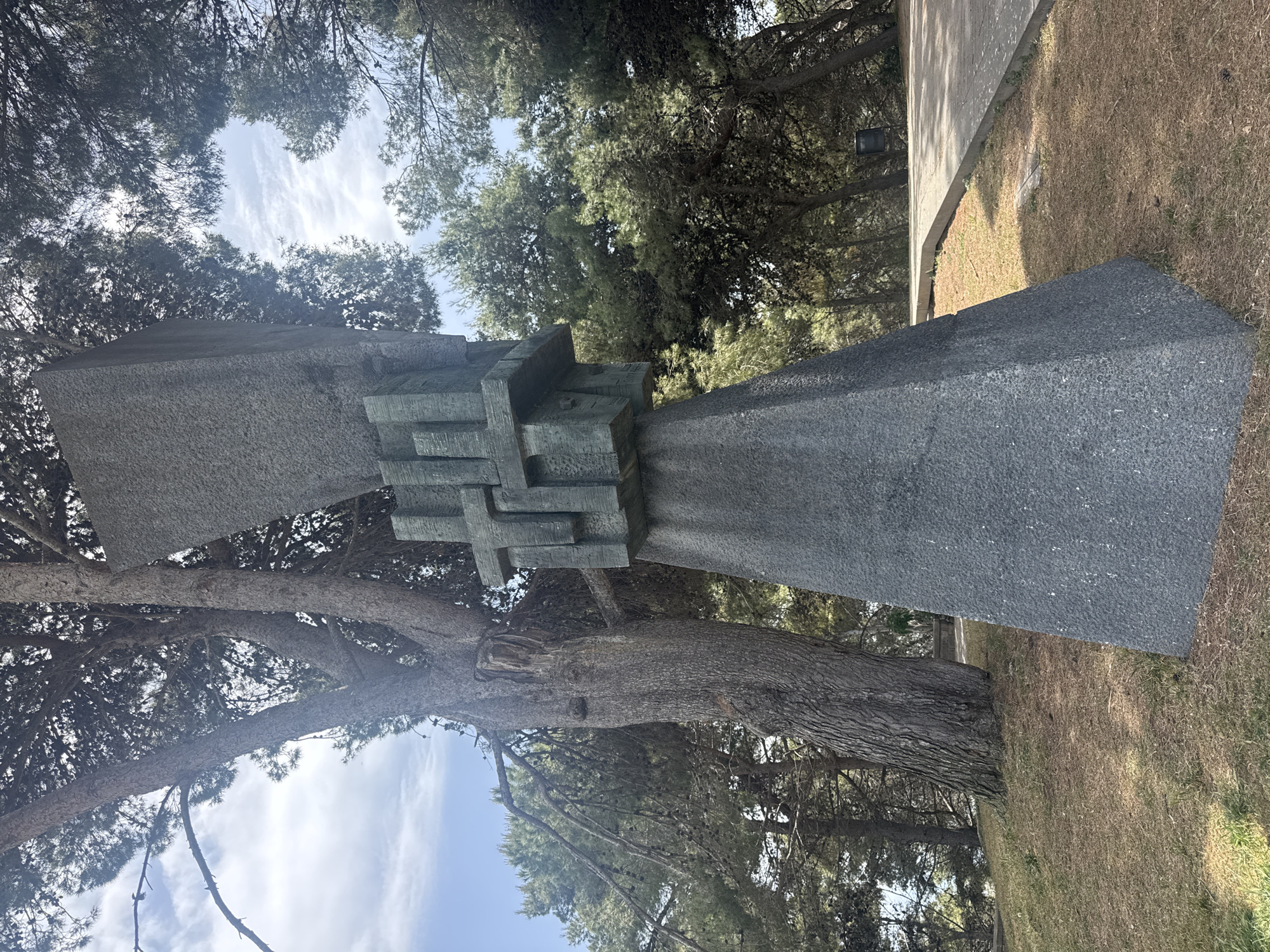 The image displays a prominent abstract stone monument set in an outdoor, park-like environment in Barcelona, Spain. The sculpture consists of two large, dark gray, speckled stone blocks, each tapering slightly towards its base. These blocks are connected in the center by a complex, intricate section of interwoven, darker-colored elements that resemble abstract crosses or interlocking rectangular pieces.
The monument is surrounded by numerous mature pine trees, characterized by their thick, textured trunks and dense, green needle foliage. The trees frame the sky above and contribute to the natural setting. The ground features dry, sparse, yellowish-brown grass and patches of exposed soil. To the right and slightly behind the sculpture, a light-colored paved path or platform is visible.
The scene is illuminated by bright daylight, suggesting late morning or early afternoon. The sky is blue with scattered, wispy white clouds, indicating clear and sunny weather. No people or activities are present, and there is no visible text on the monument or in the surrounding area. The overall impression is one of a serene, contemplative space featuring a significant piece of modern outdoor sculpture.