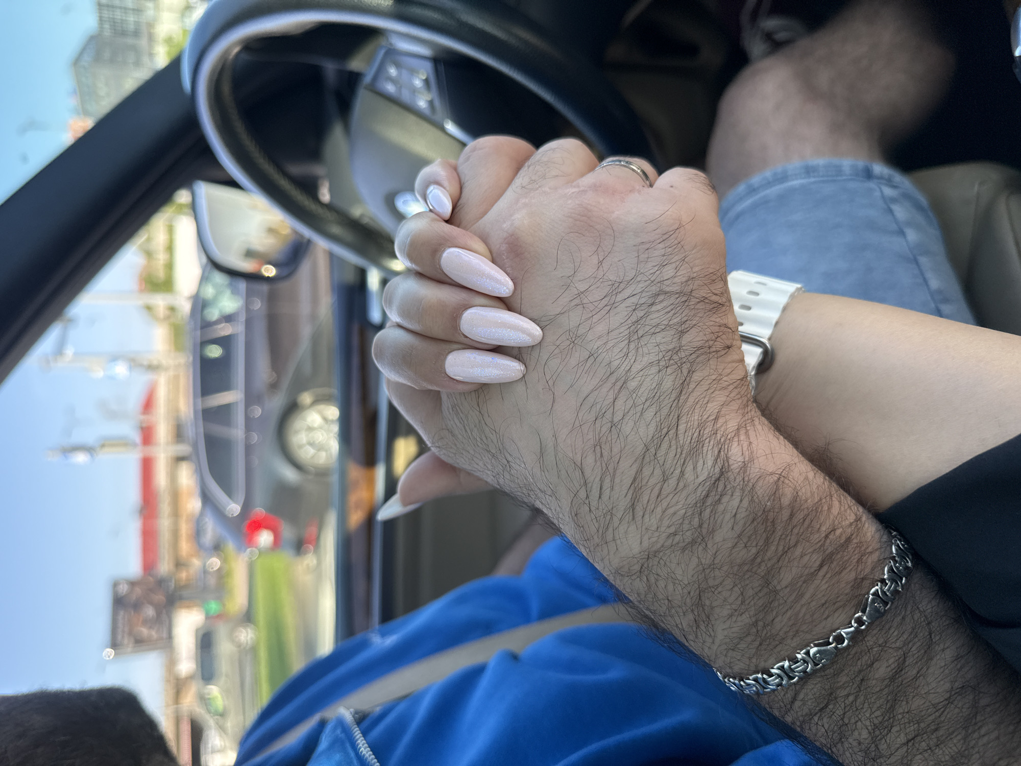 In a car, two people are holding hands. The person on the left has a blue jacket on and a silver bracelet. The person on the right has long, light pink fingernails and is wearing a white watch. They are sitting in the passenger seat and the driver's seat respectively. Through the car window, a blurred city scene is visible with buildings, trees, and other cars. The weather is sunny.