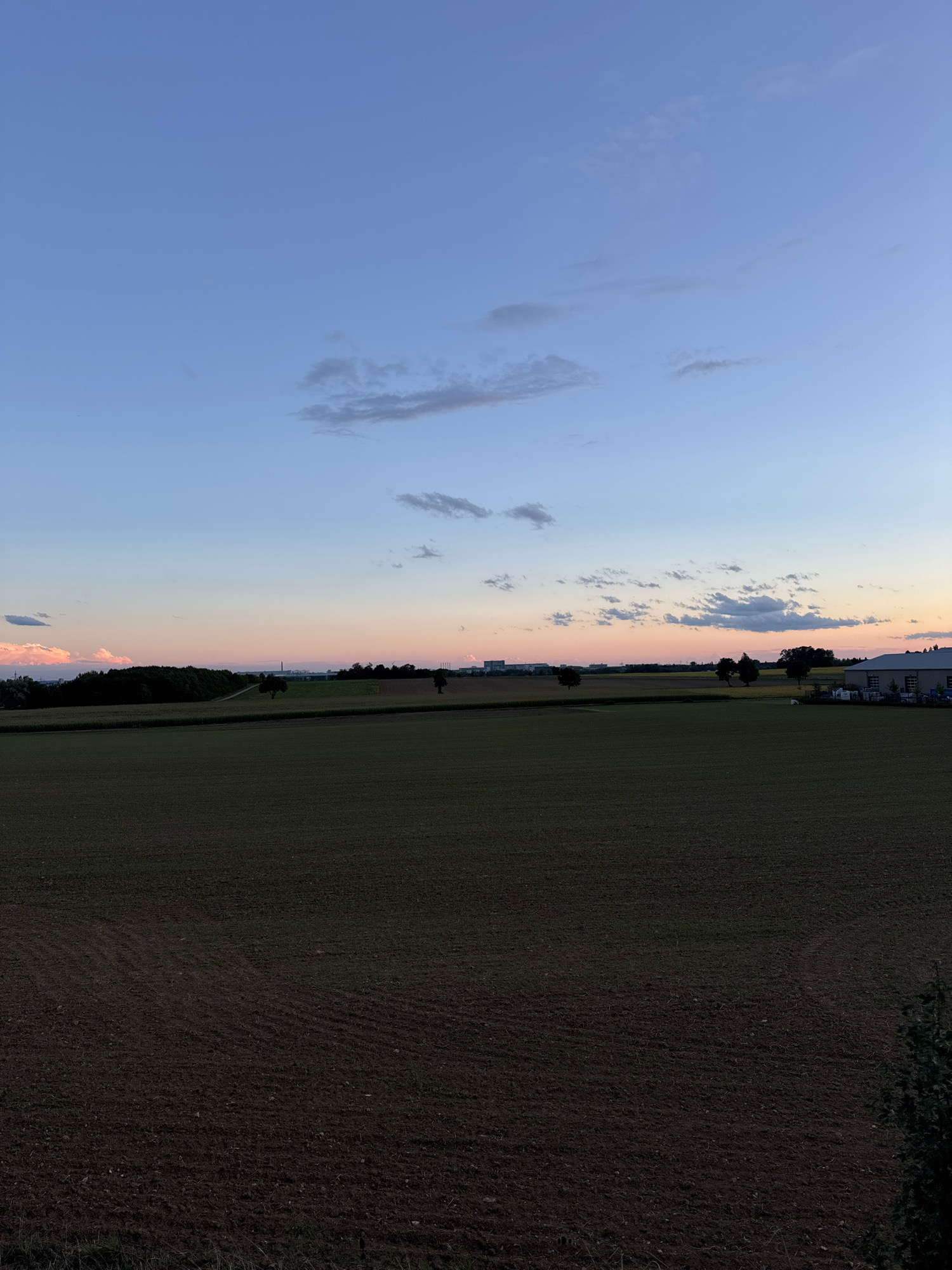 The image depicts a rural landscape at dusk, likely in Wettstetten, Germany, given the provided context. The sky transitions from a pale blue at the top to soft hues of pink and orange near the horizon. Scattered clouds are visible, some tinged with the sunset's colors.
In the middle ground, a dark green field, possibly cultivated land, stretches across the frame. Beyond this, a line of trees forms a dark silhouette against the lighter sky. Further in the distance, a low-lying industrial or commercial area with buildings and structures is visible. To the right, a modern-looking building with a grey roof and white walls is situated, possibly a warehouse or factory. The foreground is dominated by a dark, plowed field, showing the texture of the soil and faint rows from cultivation. No people are visible in the image. The overall impression is one of a tranquil, open countryside during twilight.
