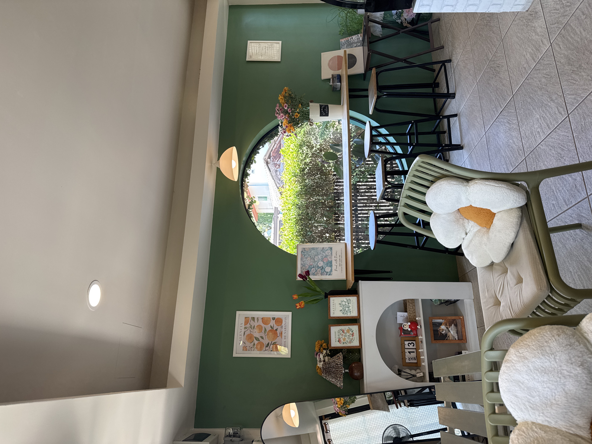 The image shows the interior of a cafe or shop in Choeng Thale, Thailand. The walls are painted green, and a large circular window provides a view of lush outdoor greenery. Several framed artworks and decorations adorn the walls. Seating includes green chairs with flower-shaped cushions, stools at a high counter, and a bench with cushions. A white cabinet is decorated with photos, decorations, and a calendar. The flooring appears to be tiled. No people are visible.