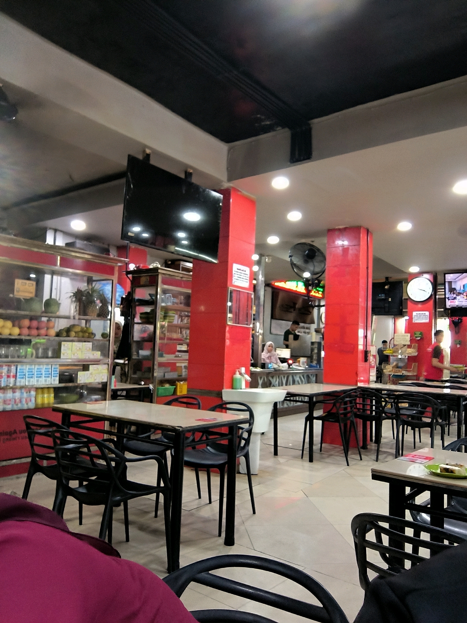 This image depicts the interior of a food establishment in Banda Aceh, Indonesia. The scene is characterized by bright lighting, with multiple ceiling lights illuminating the space. The walls are predominantly painted a vibrant red, with some areas featuring exposed ceiling structures and black acoustic tiling.
Several tables with black chairs are arranged throughout the eatery, suggesting a dining area. One table in the foreground is set with black chairs, and another table to the right has a plate with food on it. In the background, there are shelves stocked with various food items, including fruits, beverages, and packaged goods. Display cases with glass fronts are visible, showcasing the products for sale.
There are several individuals present in the establishment. In the middle ground, a person wearing a headscarf is visible behind a counter, possibly attending to customers or preparing food. To the right, another person in a red shirt appears to be working, carrying a tray. A large fan is mounted on a red pillar in the mid-ground.
Several electronic displays are mounted on the walls, including a large television screen and a clock. The flooring appears to be tiled in a light color. A sink is also visible in the mid-ground, near a red pillar. The overall impression is that of a busy, well-lit casual dining or food retail space.