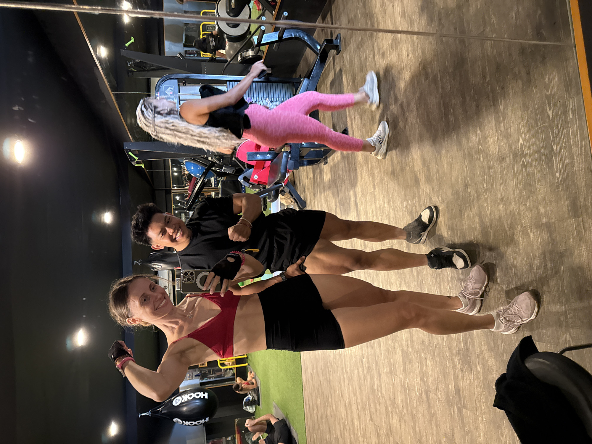 In a gym setting, a woman and a man are seen posing for a selfie. The woman, in the foreground, wears a red sports bra and black shorts, flexing her arm. Beside her stands a man in a black t-shirt and shorts. Behind them, another woman with long, gray hair is using a rowing machine. The gym's backdrop includes various workout equipment and a mirror reflecting additional gym users. The location is identified as being in the Ubud District, Indonesia.