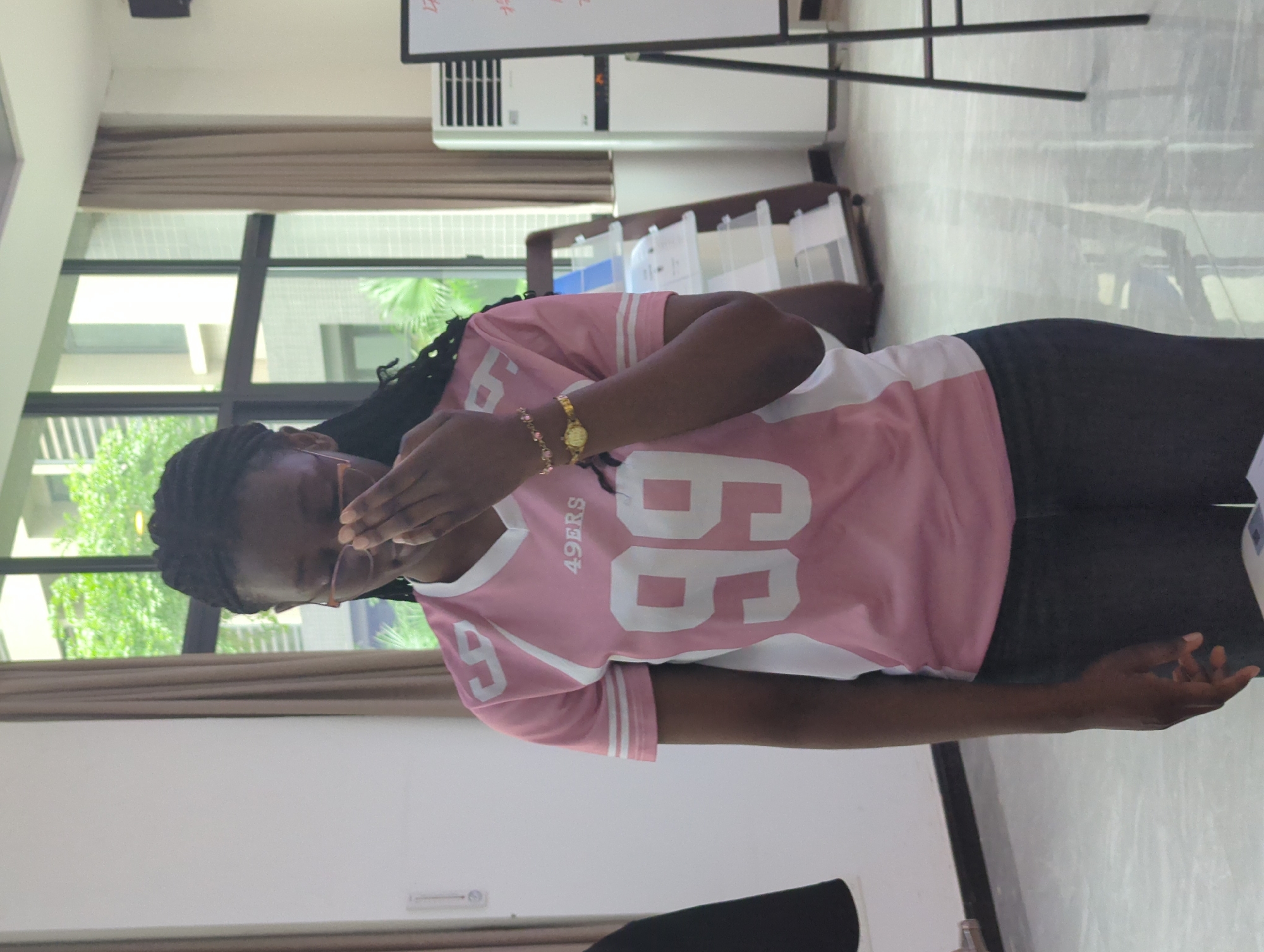 A person stands indoors. She has dark skin and wears dark-colored pants and a pink and white shirt with “49ERS” and the number “66” printed on it. She is wearing glasses, and her right hand is near her face. She has dark braids and is wearing a gold watch and gold bracelets on her left wrist.
Behind her is a window with a view of trees and buildings. A beige curtain covers part of the window. To her right is a rack holding a variety of papers or samples. In the background is a whiteboard with writing on it and an air conditioning unit. The setting appears to be in the city of Jiangjin, China, given the background architecture and vegetation. The foreground shows a portion of a black chair. The lighting is bright and diffused.