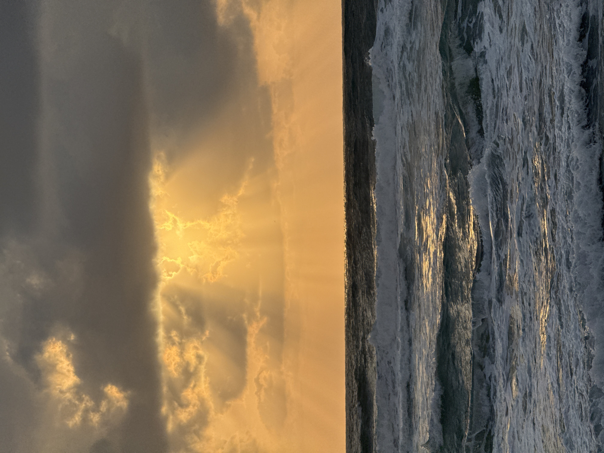 The image depicts a sunrise over the ocean in Tel-Aviv, Israel. The left side of the frame is dominated by dark, heavy clouds with golden sunlight breaking through. The right side shows the ocean, with waves crashing. The sunlight reflects off the water, creating a shimmering effect. A dark line separates the sea and sky.