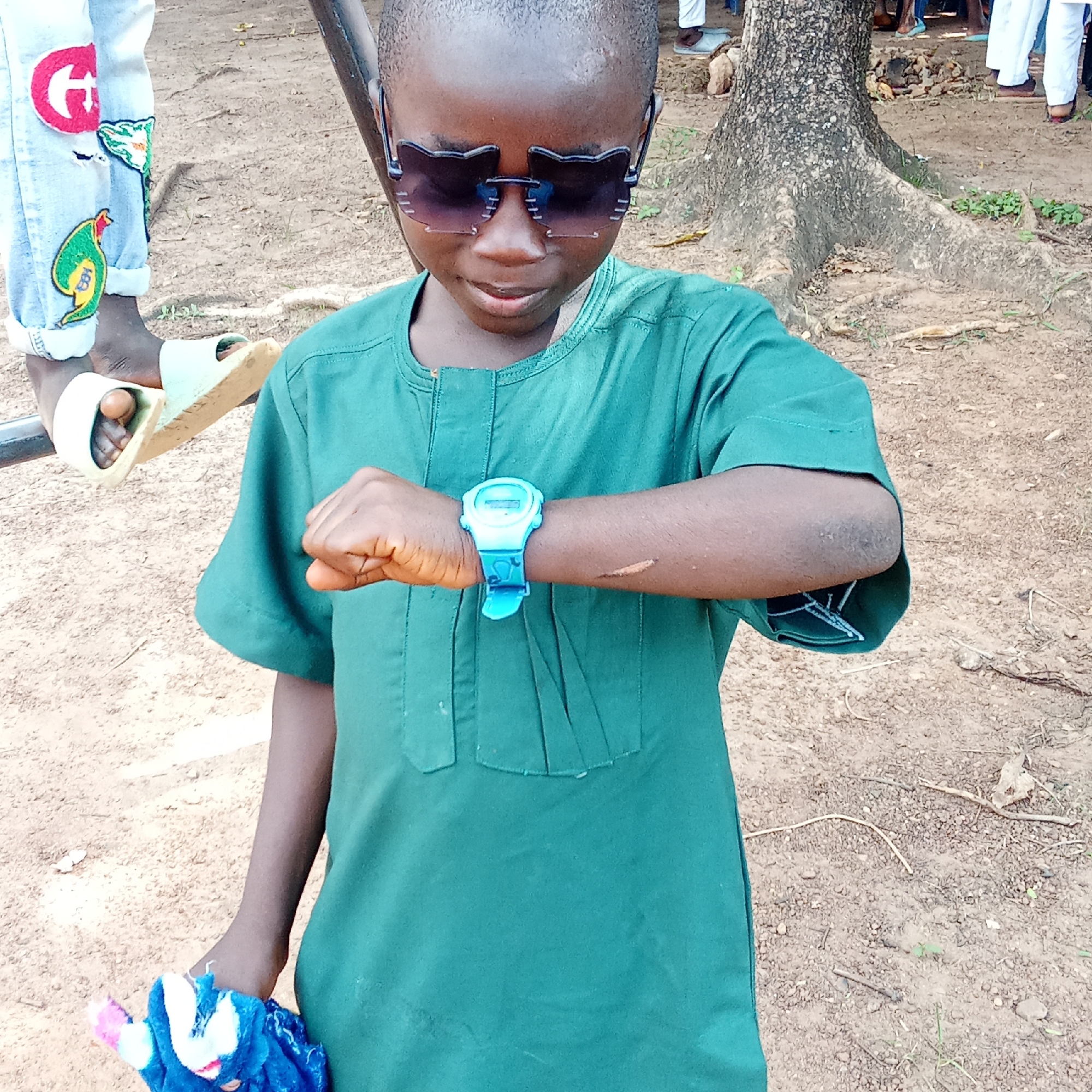 A young African male wearing an emerald green tunic and dark sunglasses stands on a dirt ground. He is looking down at a blue digital watch on his left wrist. He holds a blue and white cloth in his right hand. In the background are legs and feet of other people, a tree, and a metal bar. The location is believed to be in the city of Akwanga, Nigeria.