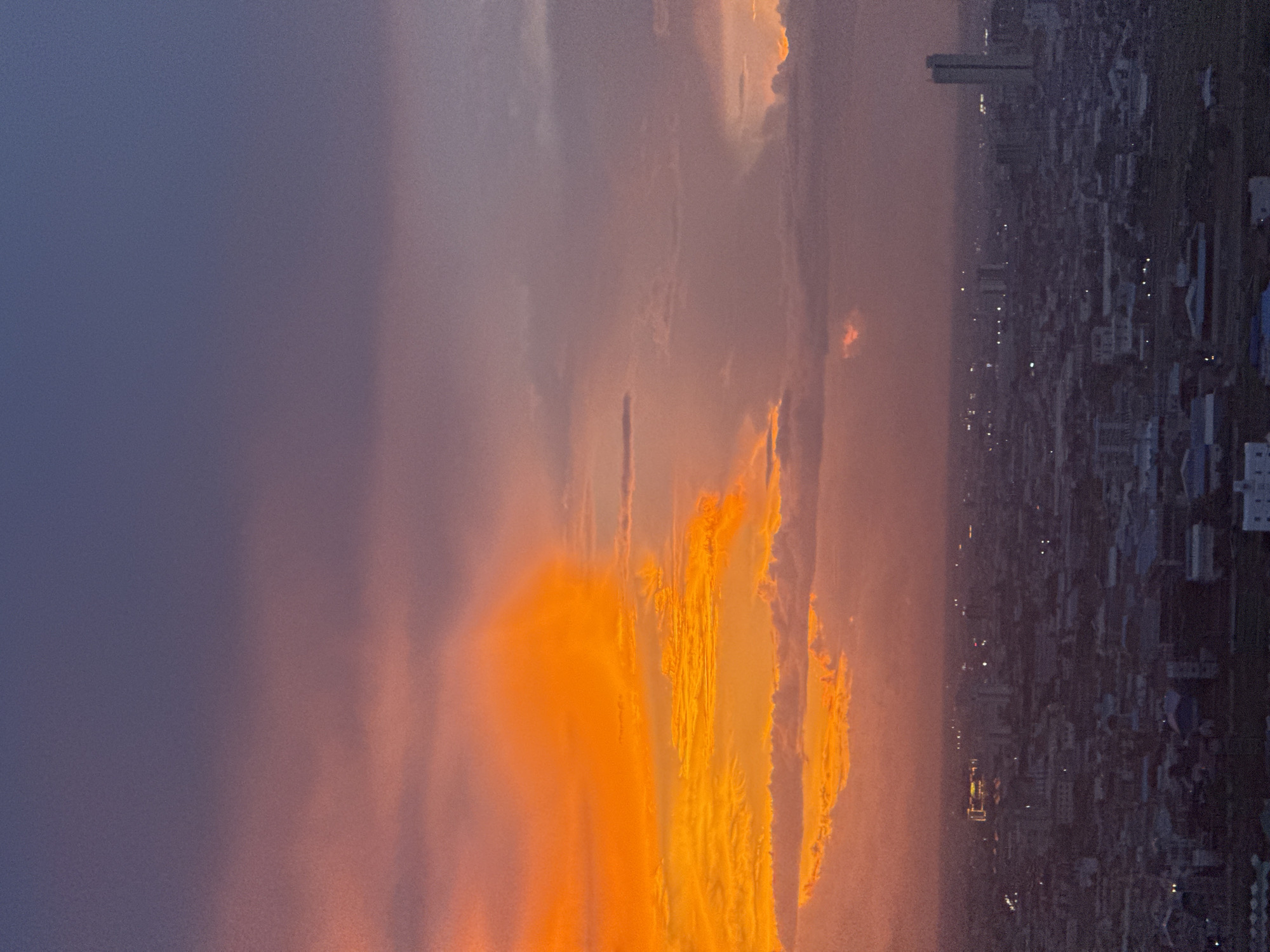 An aerial view captures a vibrant sunset over the city of Khan Mean Chey, Cambodia. The sky is a dramatic display of color, transitioning from a deep purple at the top to vivid orange and yellow hues where the sun is setting. Wispy clouds are illuminated by the low light, creating a textured effect. Below the horizon, the urban landscape of Khan Mean Chey is visible, characterized by a dense collection of buildings and structures. The city lights are beginning to appear as dusk settles, with scattered points of light indicating the ongoing activity within the city. A tall, slender building stands out on the right side of the image, a prominent landmark against the darkening skyline. The overall scene depicts the transition from day to night in an urban environment under a colorful sunset.