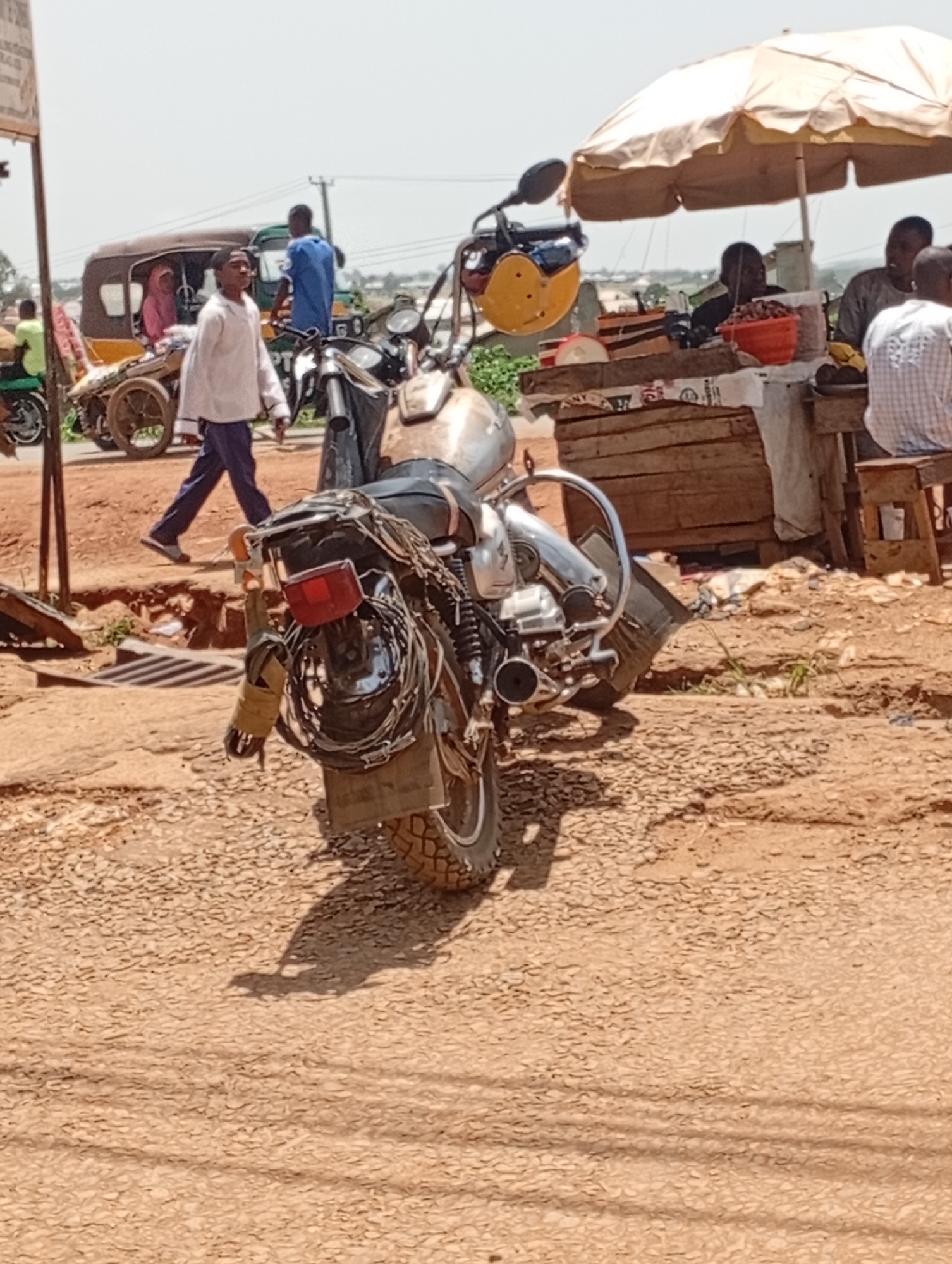 The image depicts a street scene in Rafin Par, Nigeria. In the foreground, a motorcycle is parked. Behind the motorcycle, a man in a white shirt and blue pants walks away from the camera. To his left, a yellow and brown tuk-tuk is parked, with a woman in a pink headscarf and a person in a blue shirt visible inside. Further back, under a beige umbrella, a stall is set up with people seated and goods displayed. The ground is unpaved and appears to be dusty. The overall lighting suggests a sunny day.