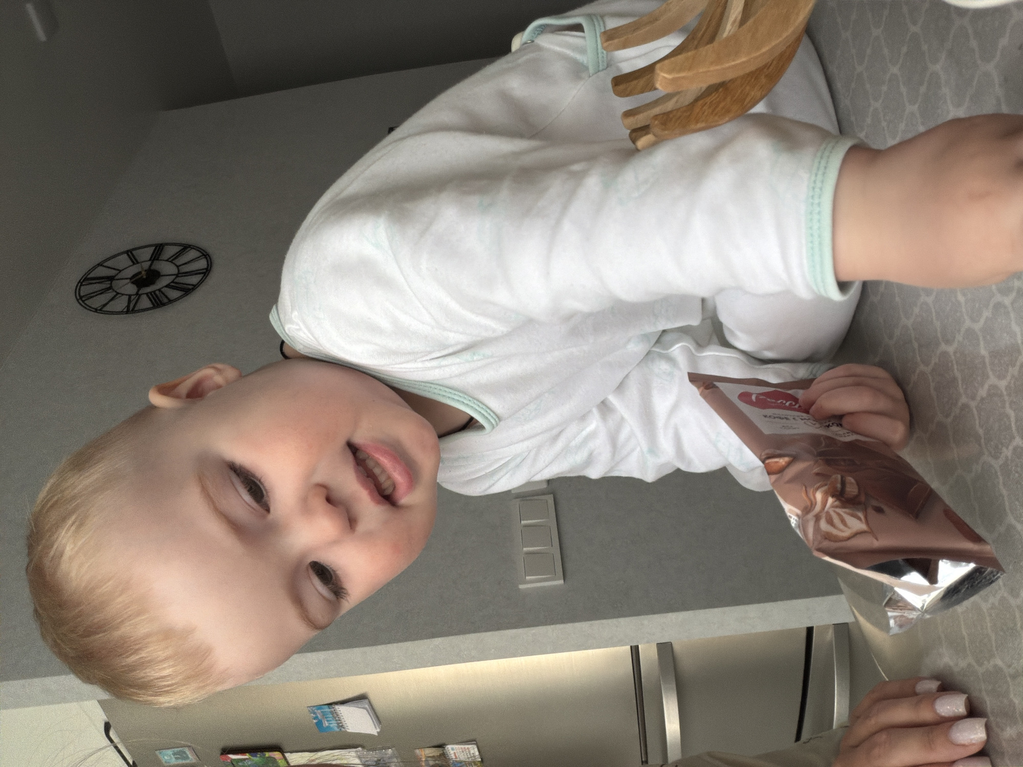 A blond-haired child wearing a white, long-sleeved garment is partially visible. The child is situated on a surface or table, holding an open package of what appears to be coffee or chocolate-flavored instant drink mix. Wooden hangers rest on the child. The environment includes a light gray wall with a light switch, a metallic refrigerator door, and wall-mounted fridge magnets and papers. A black metal wall clock is mounted above the child. There's no information confirming that this picture was taken in Votkinsk, Russia.