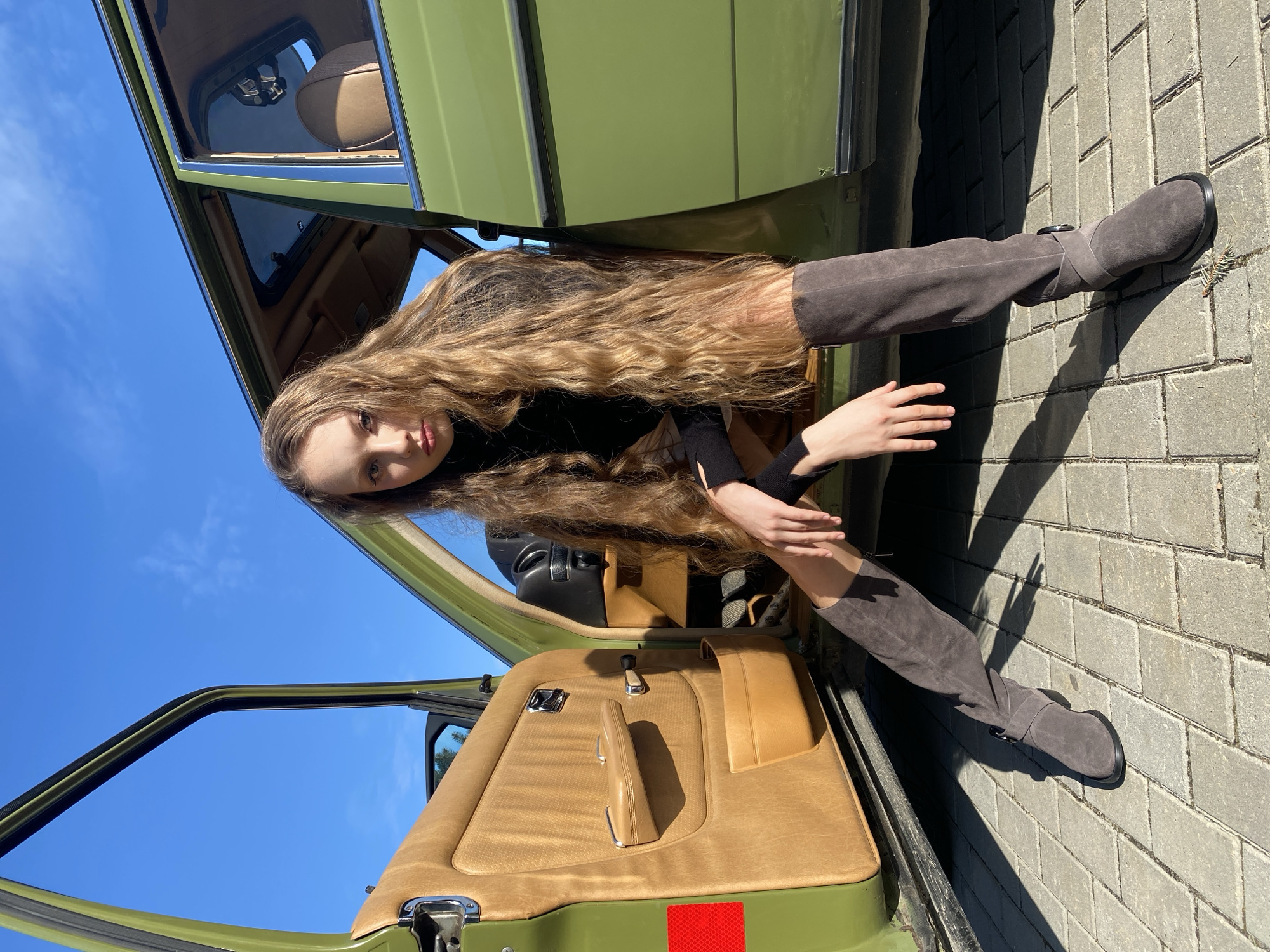 A young woman with long, wavy blonde hair is posed partially inside and outside of a vintage green car. She is wearing a black turtleneck sweater and brown suede knee-high boots. Her upper body and head are inside the car, while her legs extend out onto a brick walkway. The car's interior is tan, with an open door revealing the bright blue sky outside. The car's exterior is a muted green. A small red reflector is visible on the car door. The location cannot be confirmed as Yekaterinburg, Russia from provided image and hints.