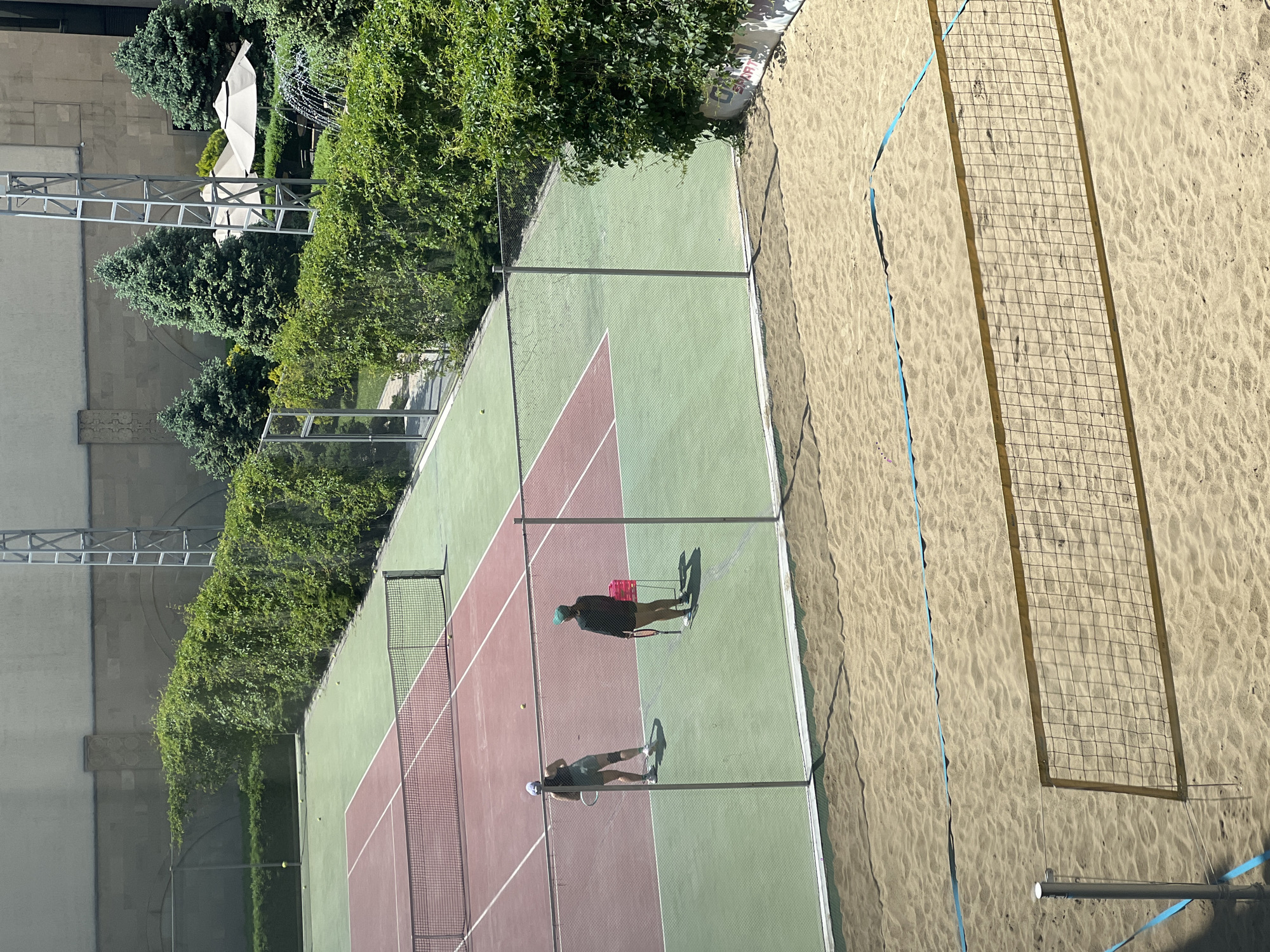 The image presents an elevated view of outdoor sports facilities, possibly located in Yerevan, Armenia. The scene includes a tennis court with green and reddish-brown surfaces, along with a sand volleyball court adjacent to it. The tennis court is enclosed by a wire fence and features players in action. A net divides the tennis court. Near the volleyball court, a net stands on the sandy surface. Vegetation, including trees and hedges, borders the facilities. In the background, structures like metal frameworks and light-colored buildings are visible. The image suggests a sunny day due to the presence of shadows.