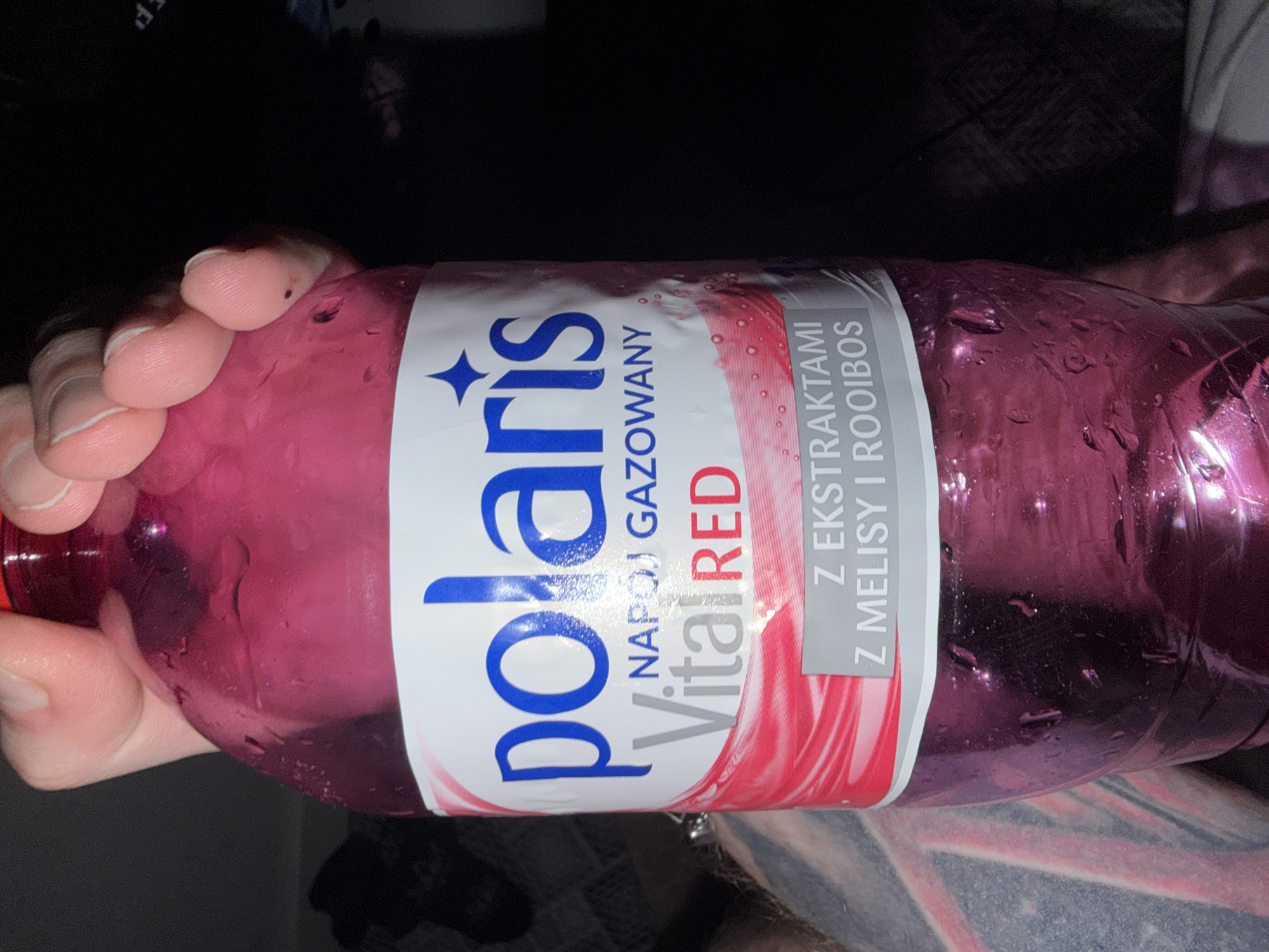The image shows a person holding a pink bottle of Polaris Vital Red flavored carbonated water. The bottle has a white label with blue lettering that reads "polaris" and "NAPÓJ GAZOWANY" which means "carbonated drink" in Polish. Below that, it says "Vital RED" and "Z EKSTRAKTAMI Z MELISY I ROOIBOS," indicating it contains extracts of lemon balm and rooibos. The bottle is condensation-covered, suggesting it is cold. The background is dark and out of focus, with what appear to be dark furnishings or clothing. The image was taken indoors. The location in Koszalin, Poland is not ascertainable from the provided image.