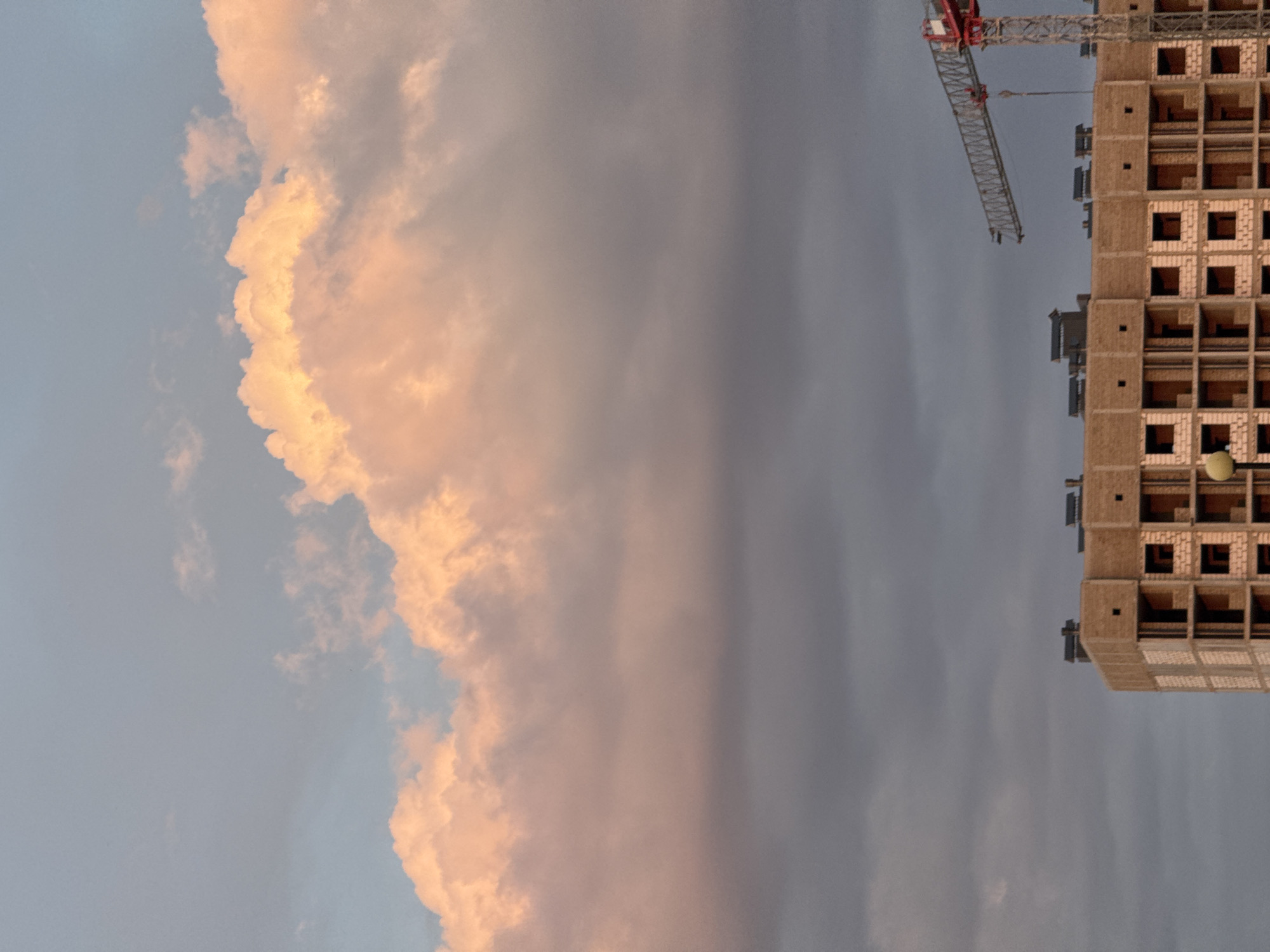 A construction site is visible in Mikhaylovsk, Russia, under a dramatic sky. The image captures the upper portion of a building under construction, characterized by exposed brickwork and window openings. A large red and grey crane is positioned to the right of the building, extending over its structure. The sky above is a mix of dark grey clouds and lighter, pink-hued clouds, suggesting either sunrise or sunset. The overall scene conveys a sense of ongoing development within the urban landscape.