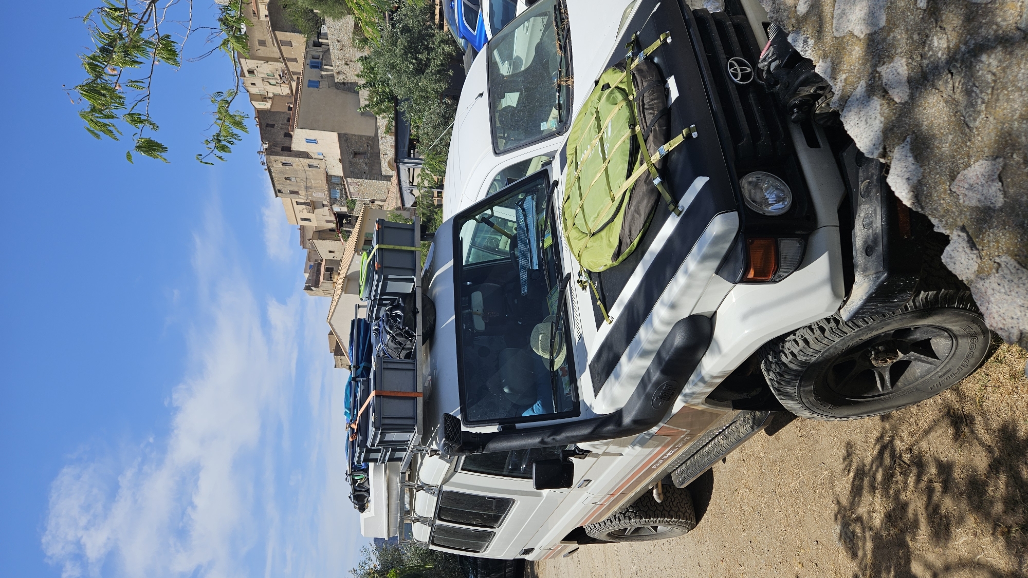 A white, four-wheel drive vehicle with a snorkel is parked on a dirt lot. The vehicle is loaded with luggage on its roof rack, including several grey storage boxes and blue bags. A green, circular object with straps is secured to the hood of the vehicle. In the background, a village with light-colored buildings is situated on a hillside under a blue sky with scattered clouds. The scene appears to be in the town of Sant'Antonino, France.