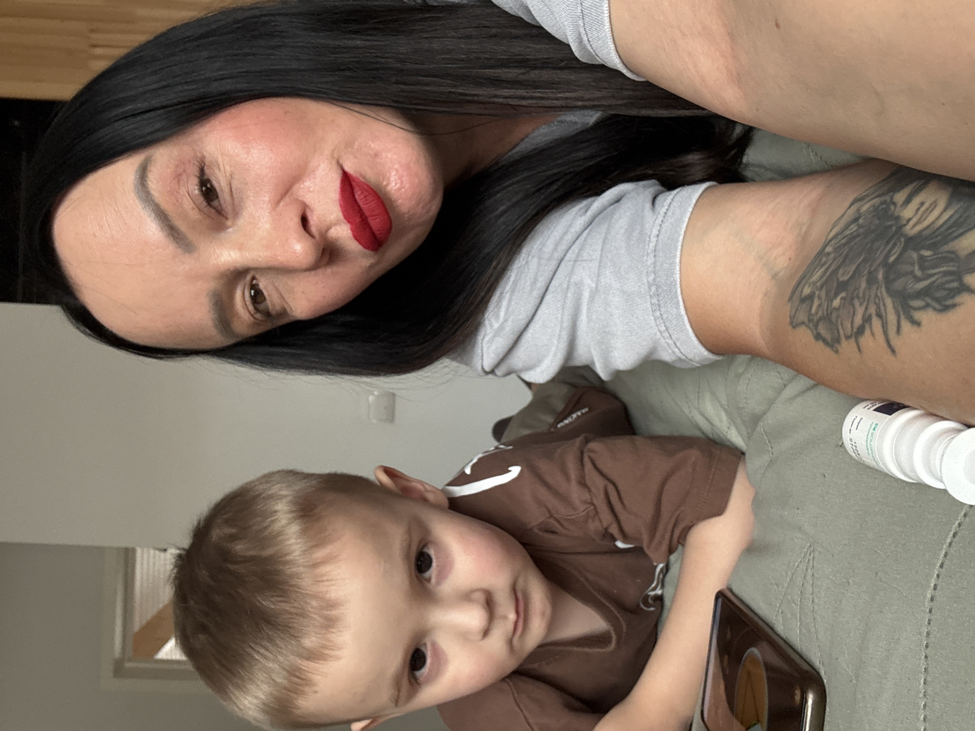 The image shows a woman and a young child indoors. The woman, who has dark hair and is wearing red lipstick, is positioned towards the upper part of the frame. Her right arm, visible in the upper right corner, has a tattoo. The young child, with light brown hair and wearing a brown shirt, is located in the lower part of the image. A cell phone is near the child. Also partially visible, in the right corner, are medicine bottles. The background includes a light colored wall. The location is assumed to be the city of Chaikovsky, Russia. No action or interaction is clear in the photo.
