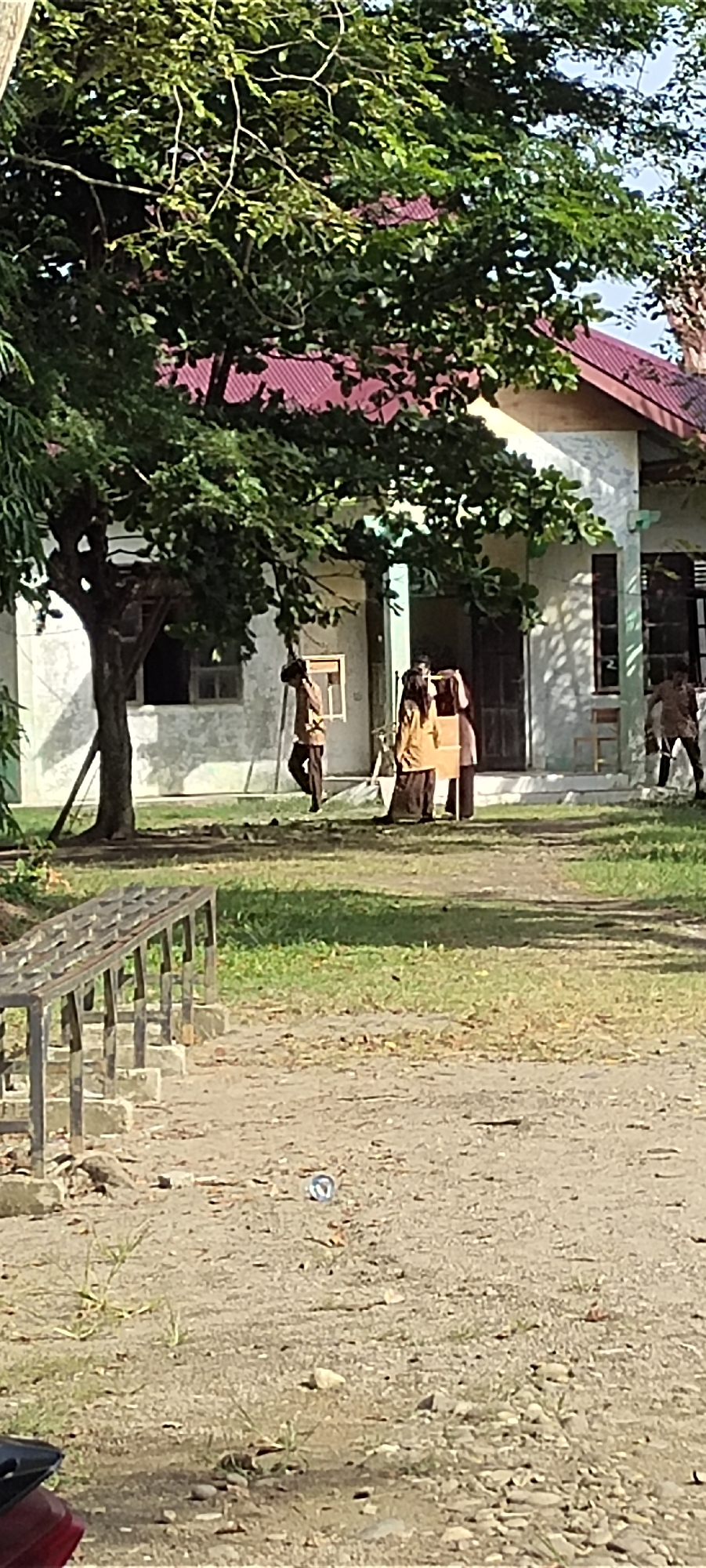 In Langnibong, Indonesia, on a sunlit day, a group of people are gathered near a light-colored building with a red-tiled roof. A large tree with dense green foliage stands to the left, casting shadows on the building and the ground. In the foreground, a metallic bench with concrete supports is visible.
Three individuals are positioned near the entrance of the building. One person, wearing a brown uniform, is walking away from the camera towards the left side of the frame, carrying an object over their shoulder. Two other individuals are standing closer together, one in a tan outfit and the other in a dark skirt and tan top, appearing to be holding a flat, rectangular object between them. Another individual in a similar brown uniform is walking towards the right, away from the building.
The ground is a mixture of dirt and sparse grass, with scattered small stones. A small, circular blue object is visible on the ground in the middle ground. The overall scene suggests an outdoor gathering or activity at a school or community building.
