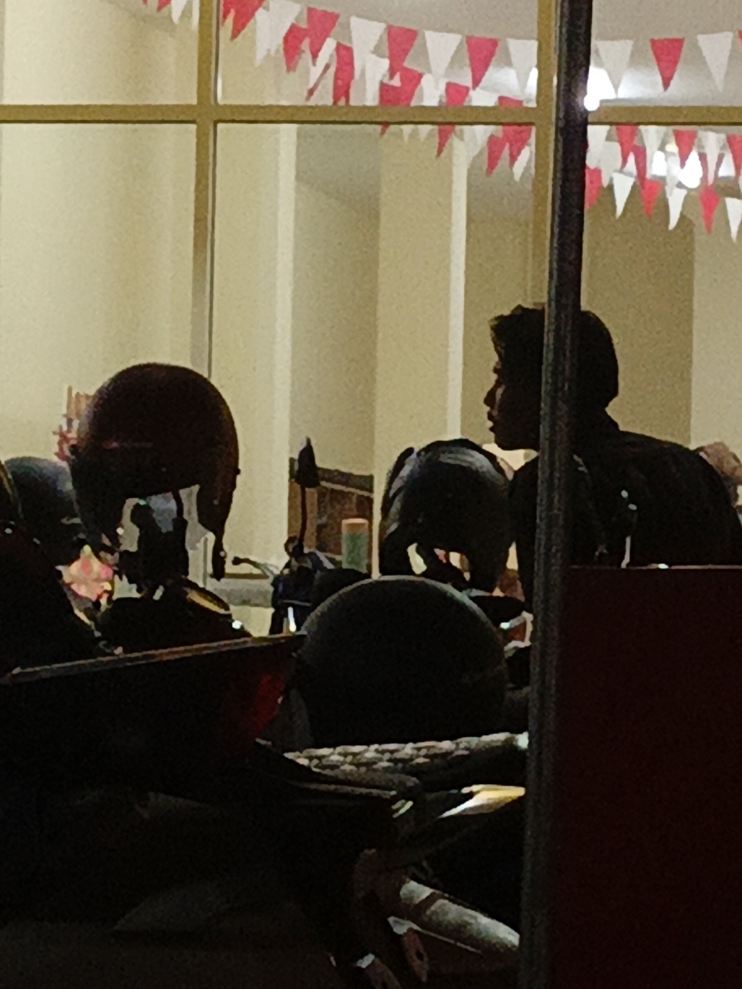In the city of Purwoharjo, Indonesia, the image shows a dimly lit interior with a person in silhouette, looking to the right, through a window. The window is adorned with red and white triangular flags. Several dark-colored motorcycle helmets are visible in the foreground, with one in the center, directly in front of the silhouetted figure. Other objects in the scene are not clearly identifiable due to the low lighting conditions and the focus being on the helmets and the silhouetted individual.