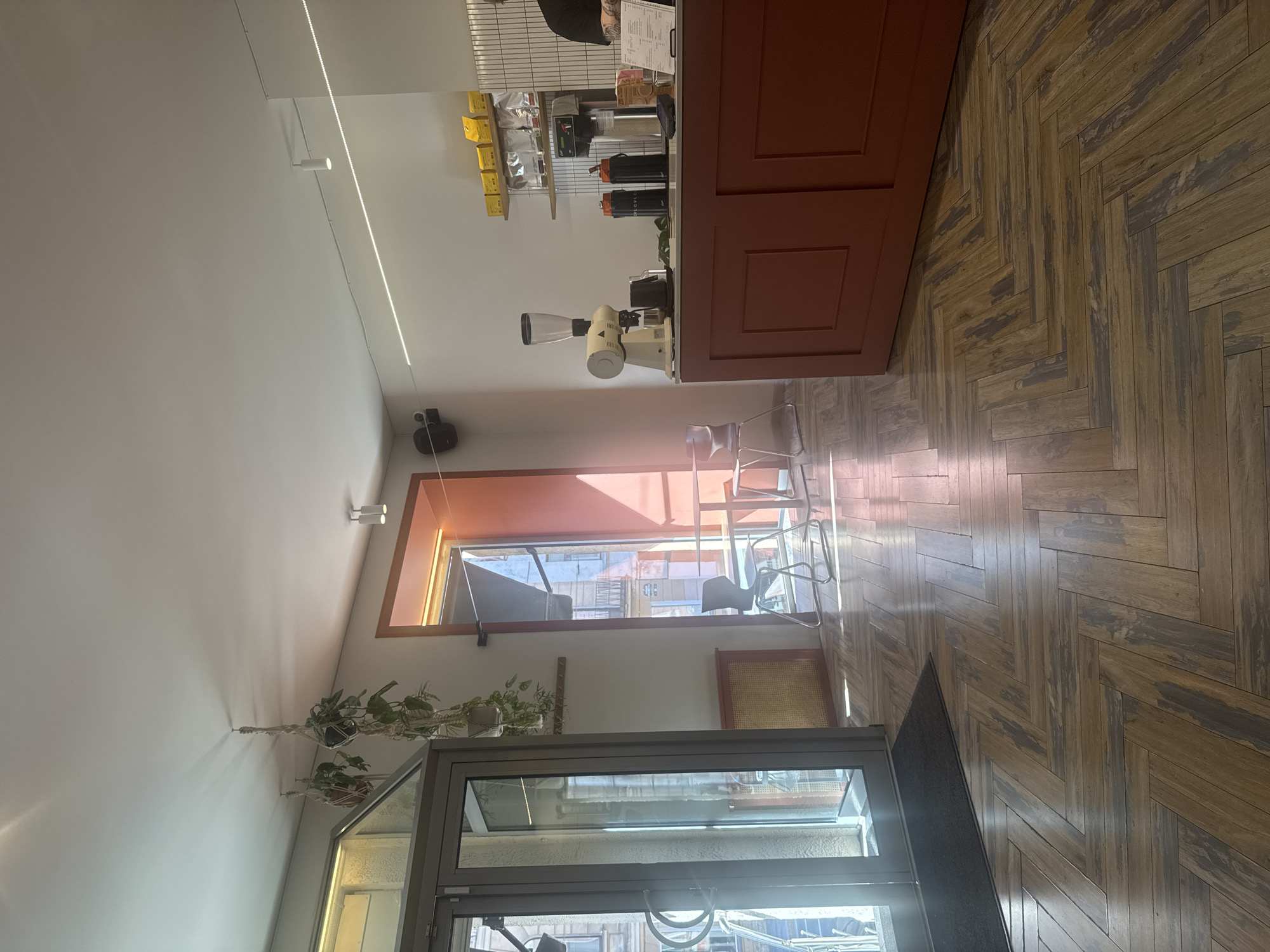The image depicts the interior of a modern coffee shop. In the foreground, a herringbone-patterned wooden floor leads towards a glass door on the left. The door has a black doormat in front of it. Sunlight streams in from the left, illuminating the floor and creating reflections.
On the right side of the image, a counter with a reddish-orange facade is visible. On top of the counter, there is a coffee grinder, a coffee machine, and two black travel mugs. Behind the counter, shelves hold yellow bags and white packages, presumably coffee beans. A menu is placed on the right side of the counter. A person, likely a barista, is partially visible behind the counter, wearing a black shirt.
To the left of the counter, a large window with an orange frame reveals an outdoor scene with a table and chairs. Two hanging plants are suspended from the ceiling, adding a touch of greenery. The walls are white, and a linear LED light runs along the ceiling. The overall atmosphere is bright and minimalist.