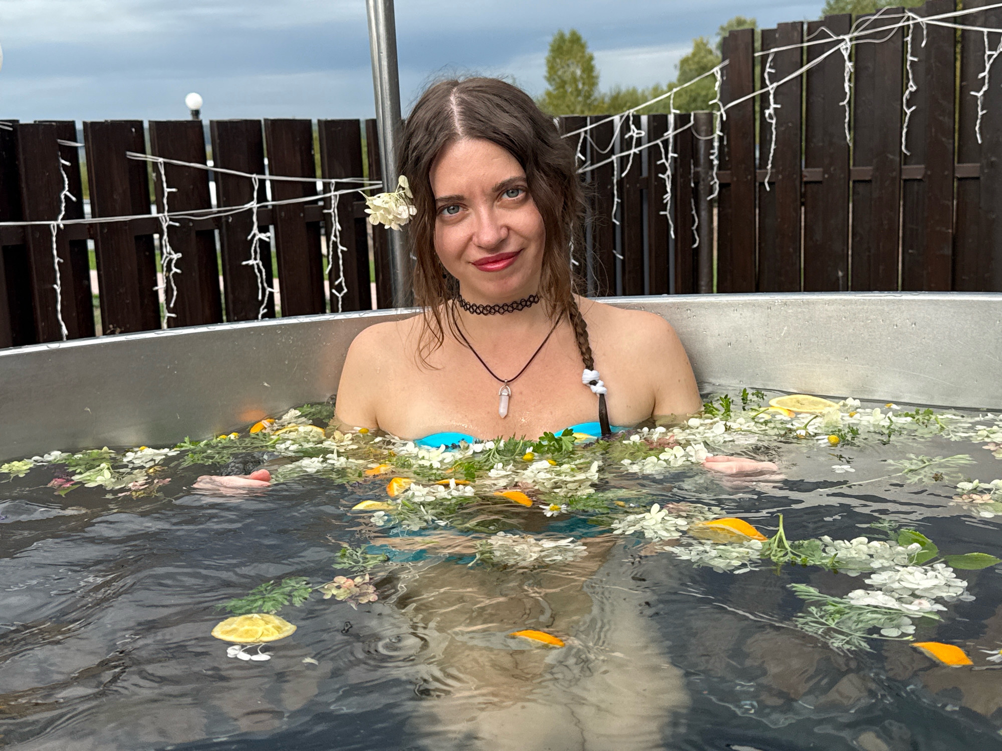 A woman with brown hair is submerged up to her shoulders in a circular metal tub filled with water, flowers, and citrus slices. She is looking at the camera and smiling. She is wearing a black choker necklace with a clear crystal pendant. Her hair is styled in loose waves with a white flower tucked behind her left ear, and a braid falling over her right shoulder. The tub is outdoors, with a wooden fence and string lights visible in the background. The sky is overcast. The location is identified as Пакли, Russia.