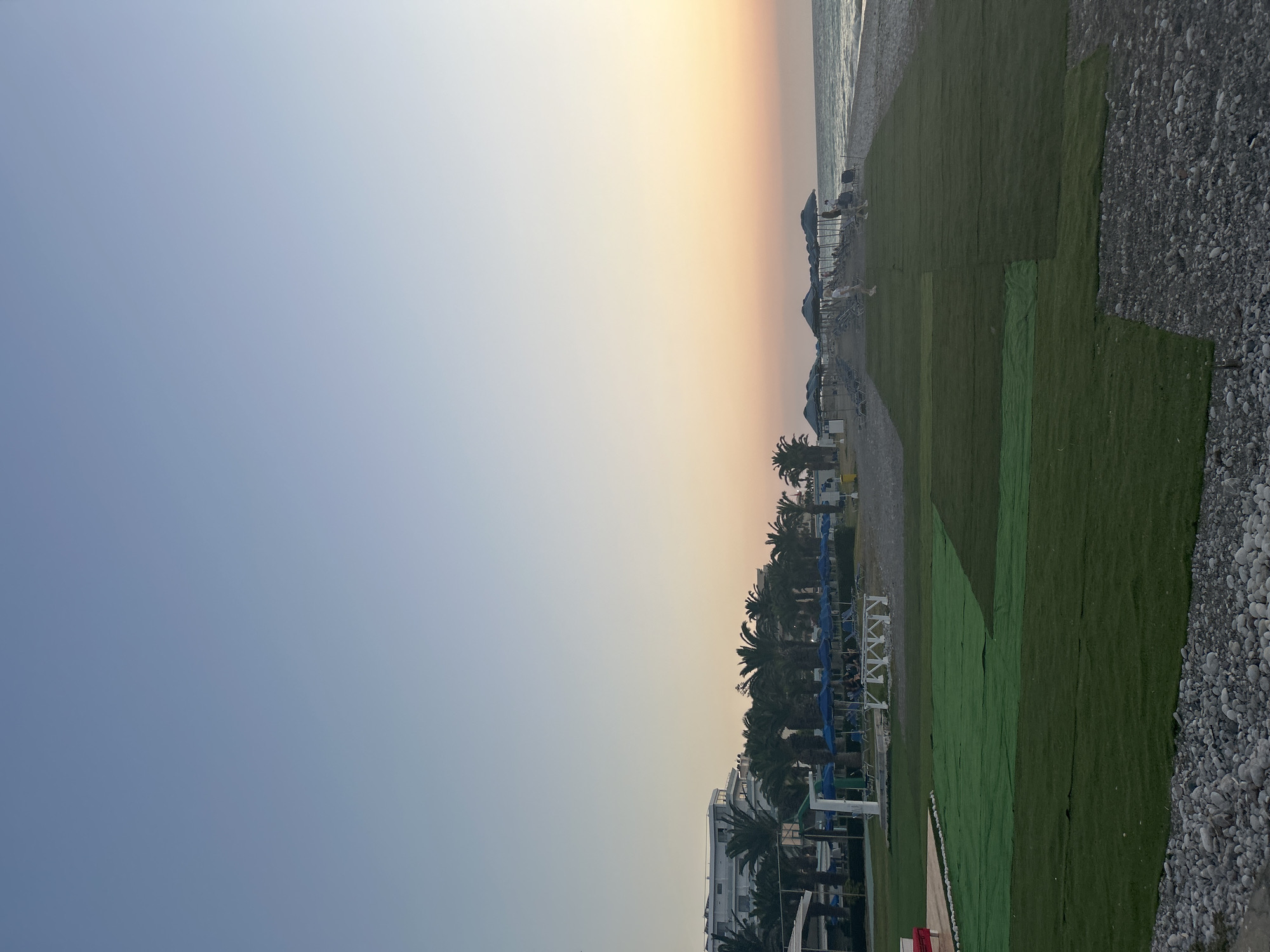This is a sideways, outdoor, vertical photograph of a beach resort in Rhodes, Greece, taken during the late afternoon or early evening. The sky is a soft gradient of blue and pale orange, suggesting either sunrise or sunset, with the sea visible as a thin line on the horizon.
The foreground on the right is a pebbly beach, transitioning to an area of artificial turf. This turf is laid in large, rectangular sections, some appearing newer and brighter green than others. Beyond the turf, a row of dark blue beach umbrellas and lounge chairs are visible, with a few people relaxing. Further inland, a line of palm trees separates the beach area from white resort buildings. Some of these buildings have balconies and modern architecture. In the background, there appear to be water slides, suggesting a family-friendly resort.
The scene evokes a sense of relaxation and leisure, typical of a vacation destination. The soft light and the presence of beach amenities indicate a peaceful end to a day. No visible text is present in the image.