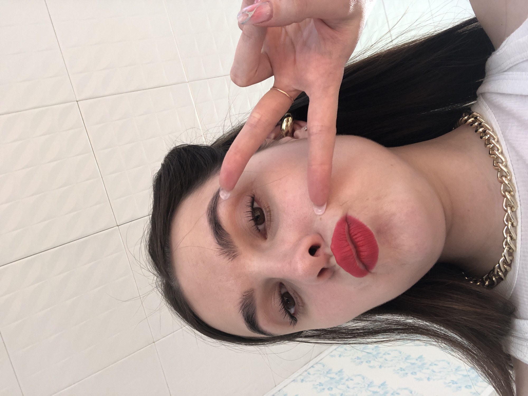 A close-up, slightly tilted portrait captures a young woman posing indoors. Her head is angled to the left, with her dark hair falling around her shoulders. She has prominent dark eyebrows and long, curled eyelashes, with her eyes looking towards the camera. Her lips are adorned with vibrant red lipstick and are slightly parted in a pouting expression.
Her right hand is raised, making a "peace" or "V" sign with two fingers extended towards her face. Her nails are manicured with a light, possibly iridescent, polish. A thin gold ring is visible on her ring finger. Around her neck, she wears a chunky gold chain necklace over a white t-shirt. A gold hoop or stud earring is visible in her right ear.
The background consists of light-colored, possibly white or off-white, square tiles with a subtle textured pattern. A small section at the bottom of the frame reveals a blue and white patterned surface, likely floor tiles or a decorative lower wall. The lighting appears to be indoor and uniform, with no specific time of day or weather discernible. There is no visible text. The location context provided is Noviy, Russia, though the image itself offers no specific visual cues to confirm this.