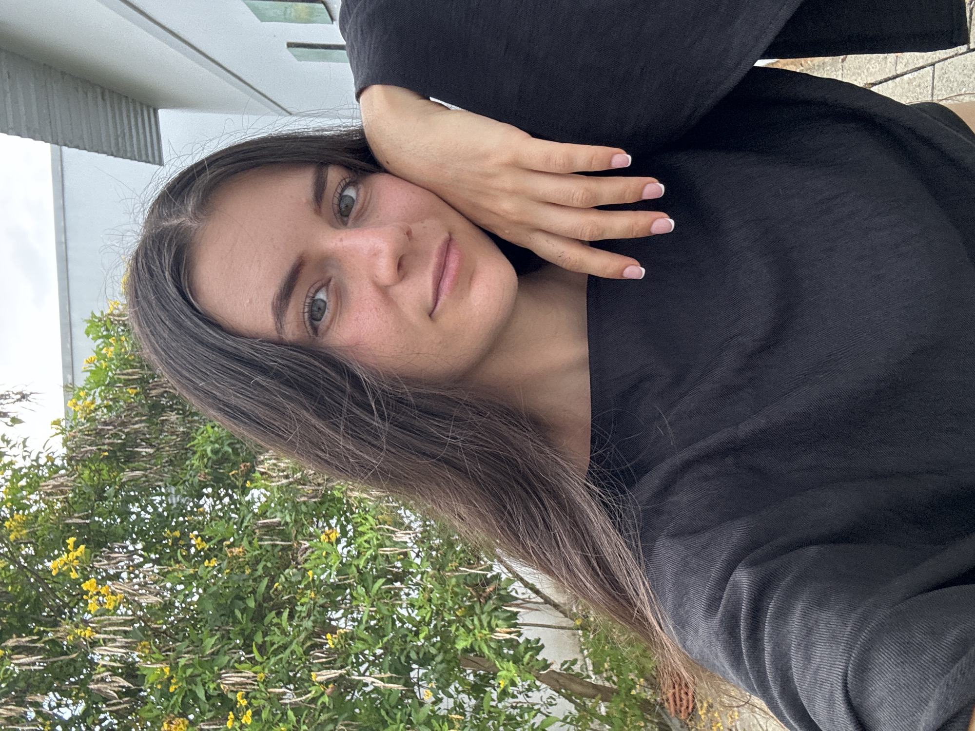 A young woman with brown hair, green eyes, and light skin is posed in a selfie-style photo. Her hand is resting on her face. She is wearing a black shirt. Behind her, there is a tree with yellow flowers and a building with a corrugated metal roof. Based on the context, this photograph could have been taken in Mũi Né, Vietnam, however, there is nothing specific in the image to confirm this location.