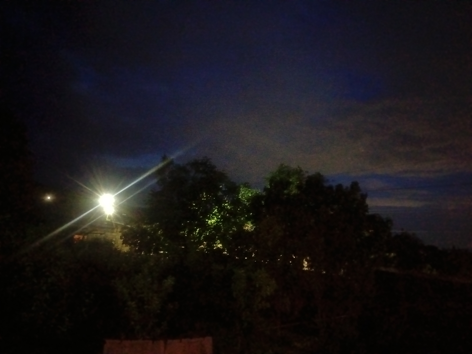 This nocturnal image captures a quiet outdoor scene in Nor Geghi, Armenia. The dark sky, a blend of deep blue and black, is partially obscured by clouds, which are more visible and appear lighter towards the right and upper right of the frame. A dominant artificial light source, resembling a street lamp, is positioned on the left side, emitting strong, starburst-like rays that cut across the darkness. Above and slightly to the left of this main light, a smaller, circular, fainter light source is present, possibly the moon or another distant light.
Dense foliage and trees occupy the midground and foreground, appearing mostly as dark silhouettes against the night sky. Some of the leaves in the immediate vicinity of the artificial light are subtly illuminated, revealing touches of green. Faint outlines of structures or buildings can be glimpsed behind the trees, benefiting from the light scatter. At the very bottom of the image, a horizontal reddish-brown strip suggests a wall or fence. The photograph conveys a serene, dimly lit landscape at night. There is no discernible activity taking place, and no visible text.