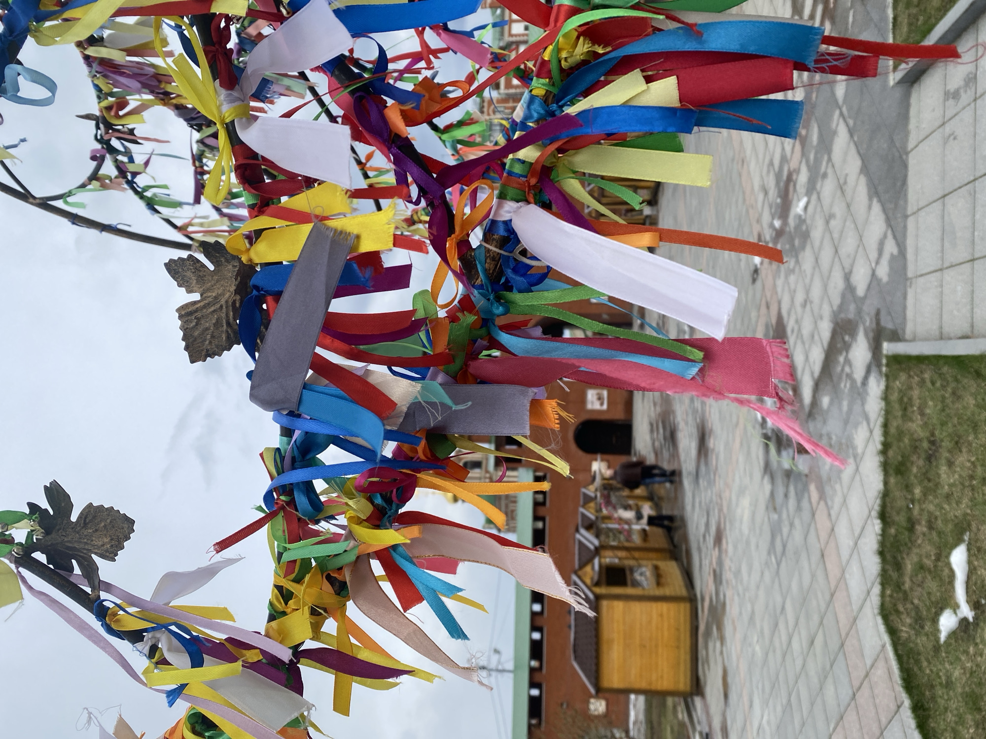 A close-up shot shows a "wishing tree" or a tree of ribbons in the city of Yoshkar-Ola, Russia. The tree branches are adorned with numerous colorful ribbons tied around them. The ribbons are in various shades including red, blue, yellow, white, and green. The tree is located in an outdoor space with a background that reveals a building constructed in a historical style. Behind the tree and ribbons, people are visible walking. The ground appears to be paved with gray tiles. To the right of the frame, there's a patch of green grass with some snow present. The sky in the background is overcast.