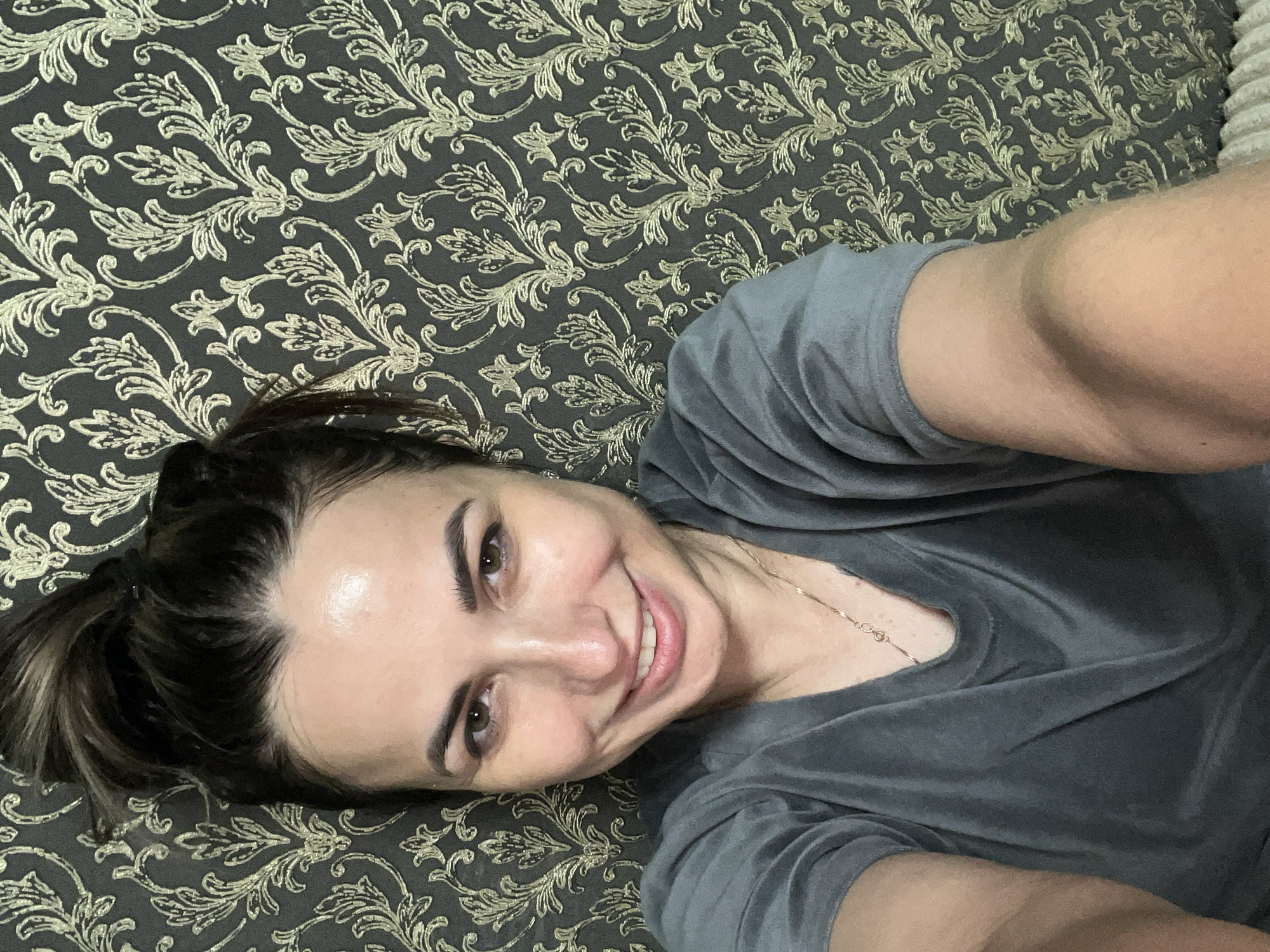 A woman with her hair in a bun is taking a selfie against a dark wallpaper with a gold floral pattern. She is wearing a grey t-shirt and a necklace. She is smiling at the camera. The image appears to be taken indoors.