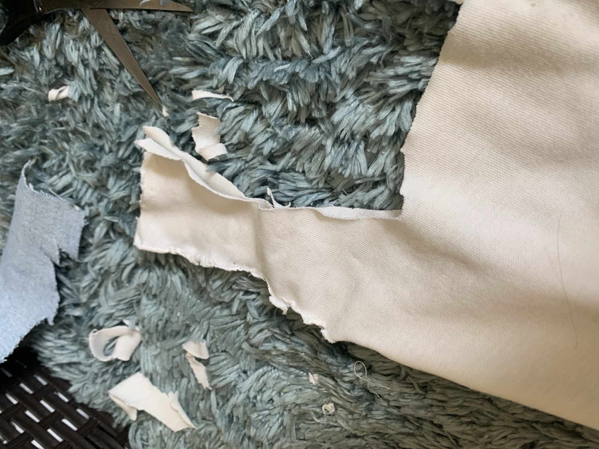 The image depicts a close-up view of fabric scraps scattered on a blue shag rug. A pair of scissors is partially visible in the upper left corner. The scraps are primarily white, with a smaller piece of light blue fabric also present. The background is dominated by the texture of the shag rug. There is no evidence of the image being taken in Vozdovac Urban Municipality, Serbia.